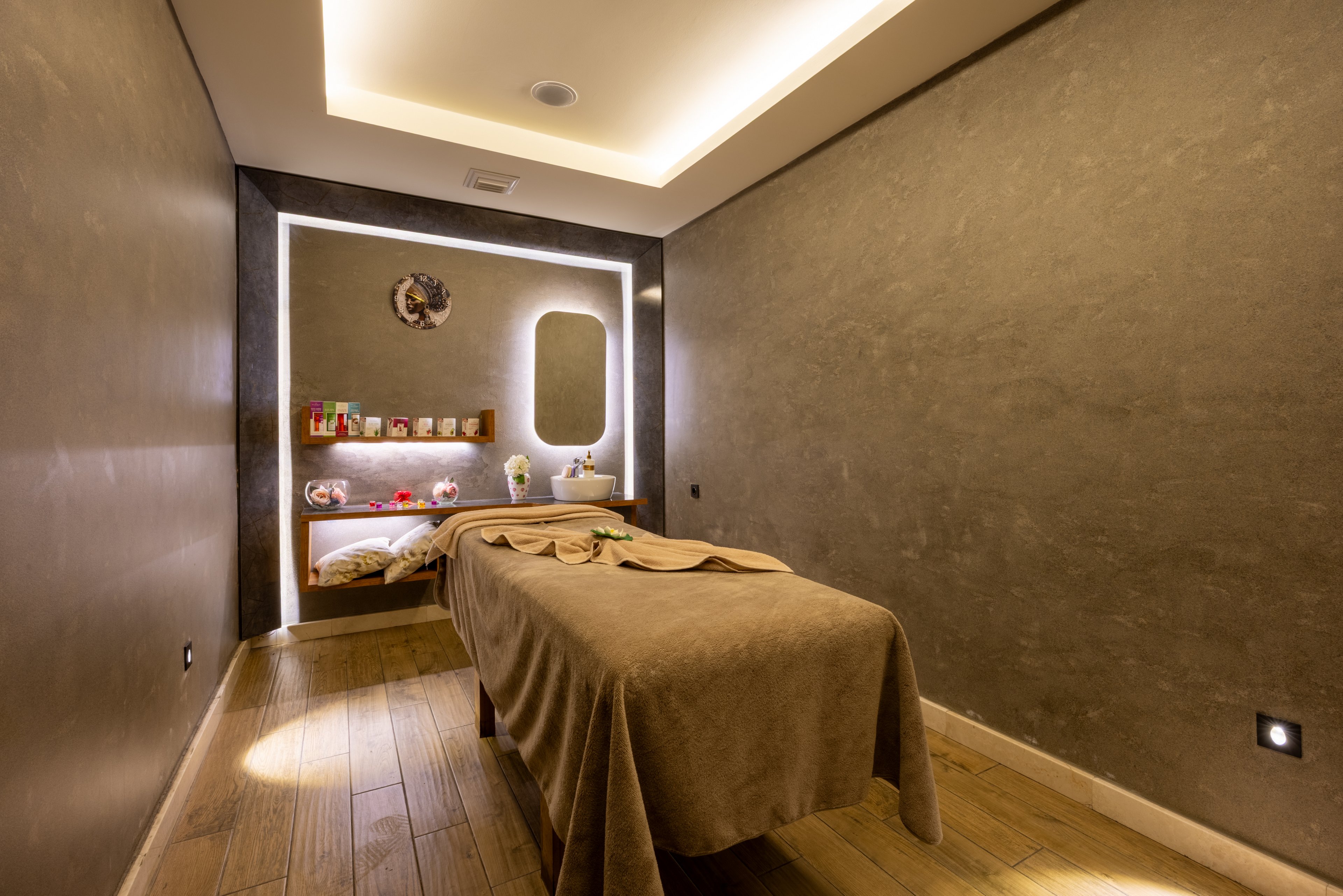 A contemporary spa treatment room features a single massage table covered with a beige sheet. The setting is tranquil, with soft, warm lighting and minimalist decor. Shelves with various spa products and a backlit mirror add to the relaxing atmosphere. The wooden floor and neutral wall tones enhance the calming mood. No visible people or text are present in the image.