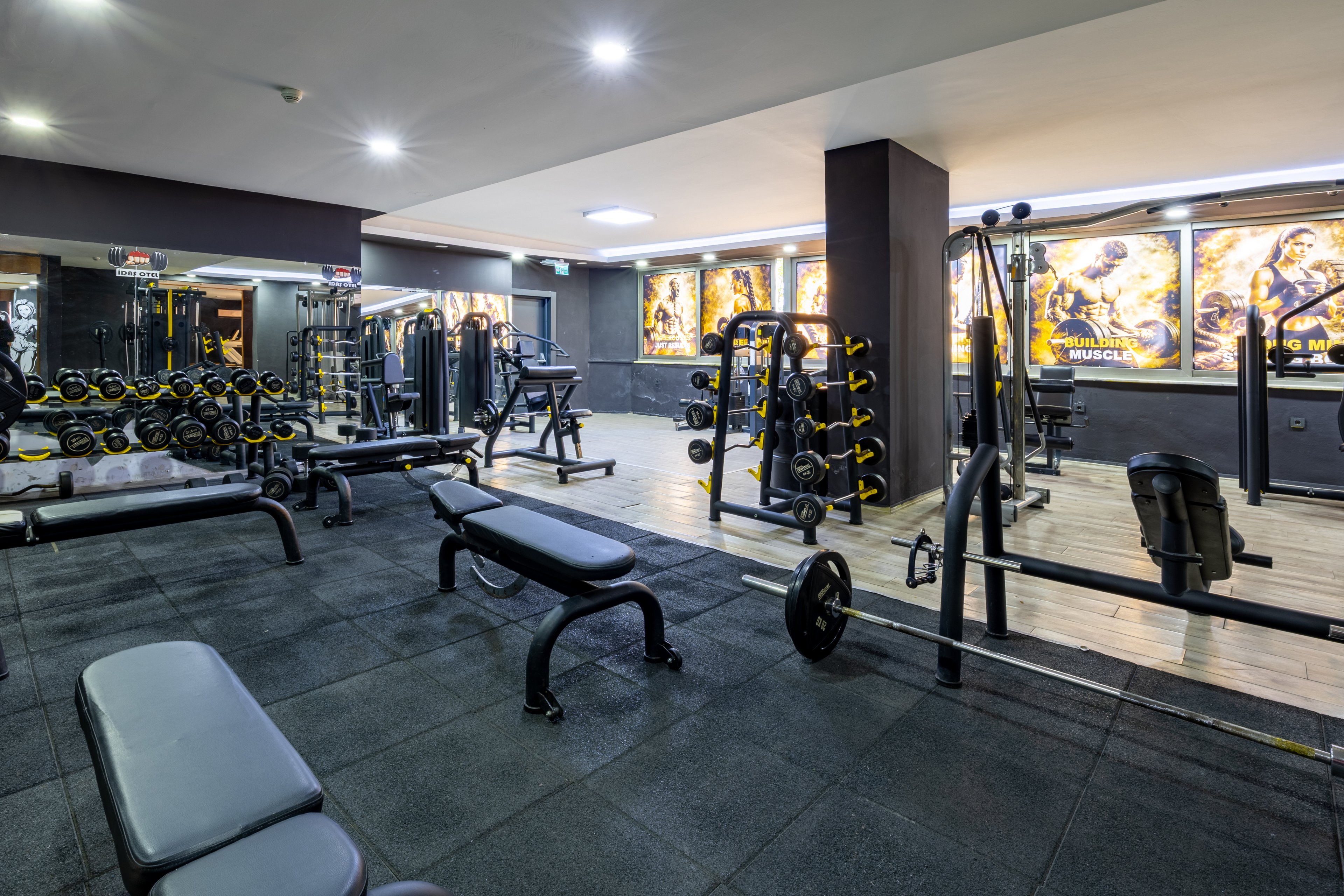 A spacious gym features various weight training machines, benches, and free weights arranged neatly. The setting is well-lit with motivational posters on the walls, creating an energetic and focused atmosphere. The color palette includes dark flooring and neutral walls, with bright accents from the posters. No people are present, highlighting the equipment and clean environment.