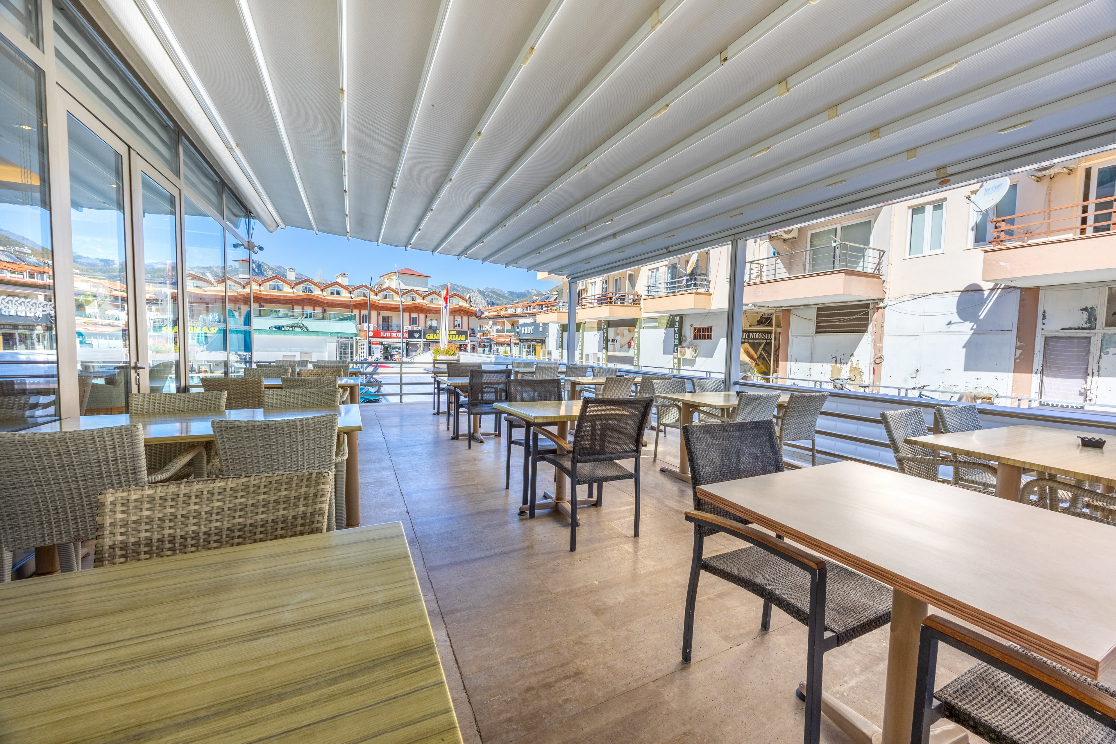 A spacious outdoor cafe terrace features neatly arranged wooden tables and wicker chairs under a white retractable awning. The setting is bright and airy, with sunlight streaming in and a view of surrounding buildings and a pool area. The atmosphere is calm and inviting, ideal for casual dining or relaxation. No people are present, emphasizing the tranquil and clean environment.