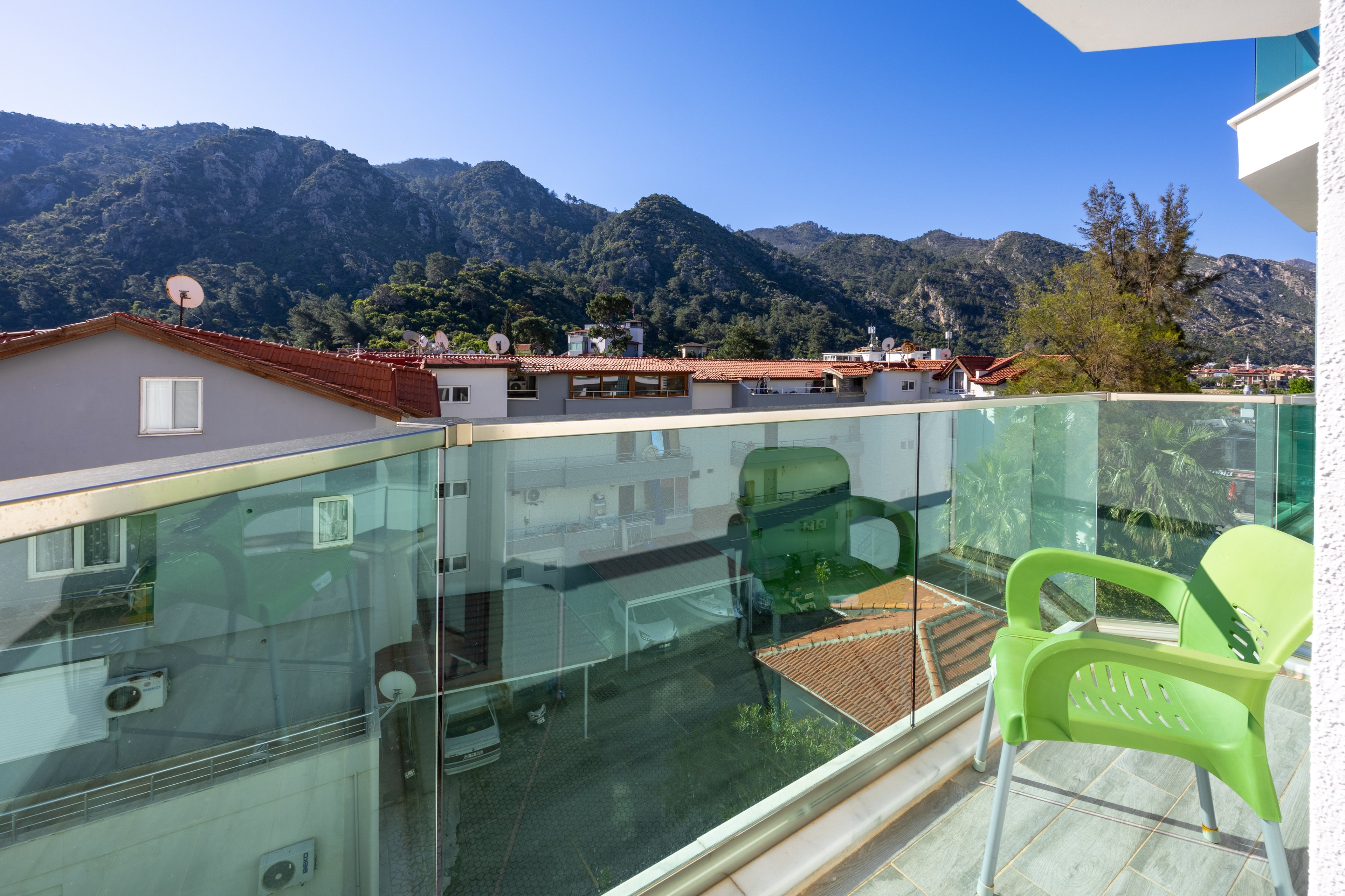 A modern balcony with glass railing features two green plastic chairs, offering a clear view of nearby rooftops and lush mountains. The setting is bright and sunny, with a vivid blue sky and strong natural light. The scene conveys a relaxed, inviting atmosphere, ideal for leisure or enjoying the outdoors. No visible text or numbers are present in the image.