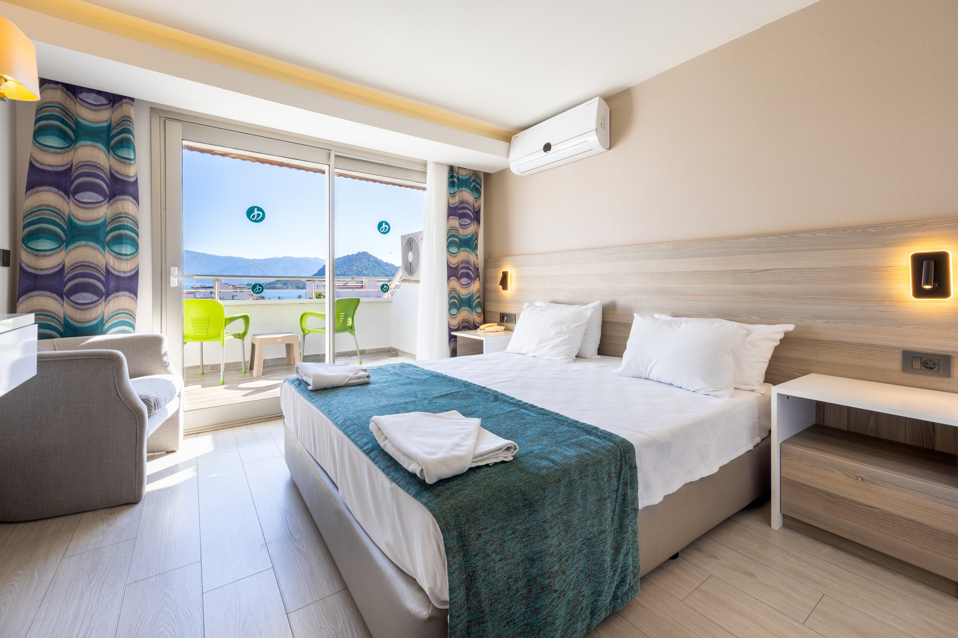 A bright, contemporary hotel room features a double bed with teal accents, bedside tables, and a comfortable armchair. Large glass doors open to a balcony with green chairs, offering a scenic view of mountains and blue sky. The room is decorated in neutral tones with patterned curtains and includes an air conditioning unit above the bed. Natural light fills the space, creating a welcoming and relaxing atmosphere. No visible text or numbers are present in the image.