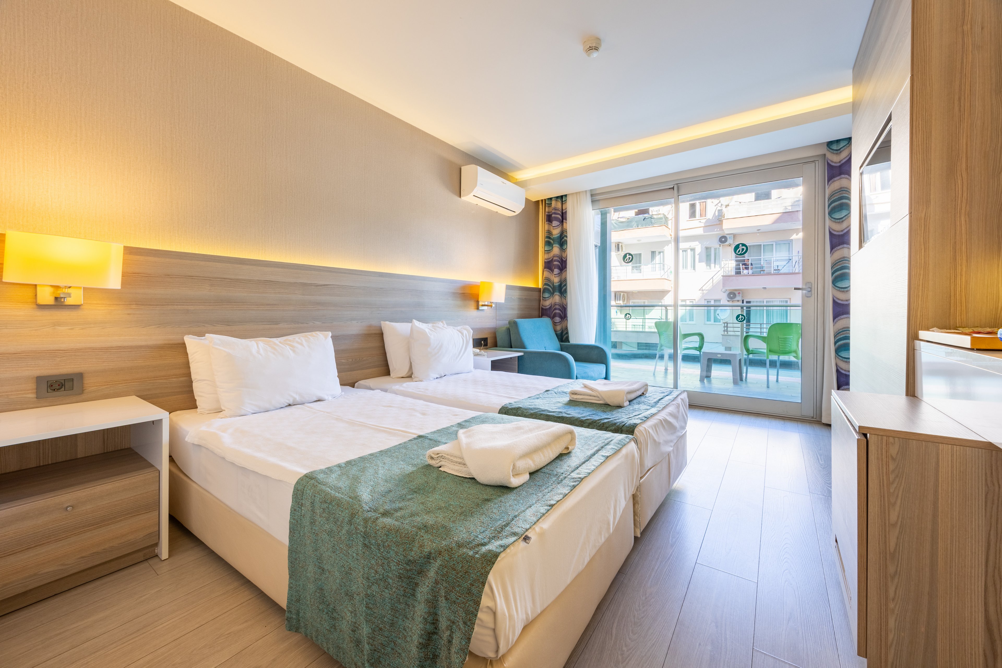 A bright, contemporary hotel room features two neatly made twin beds with teal runners and folded towels. The space is illuminated by warm wall lights and natural daylight from a large glass door leading to a furnished balcony. The room includes a small nightstand, a blue armchair, and a view of neighboring buildings. The overall palette is neutral with teal accents, creating a fresh and inviting atmosphere. No visible text or numbers are present in the image.