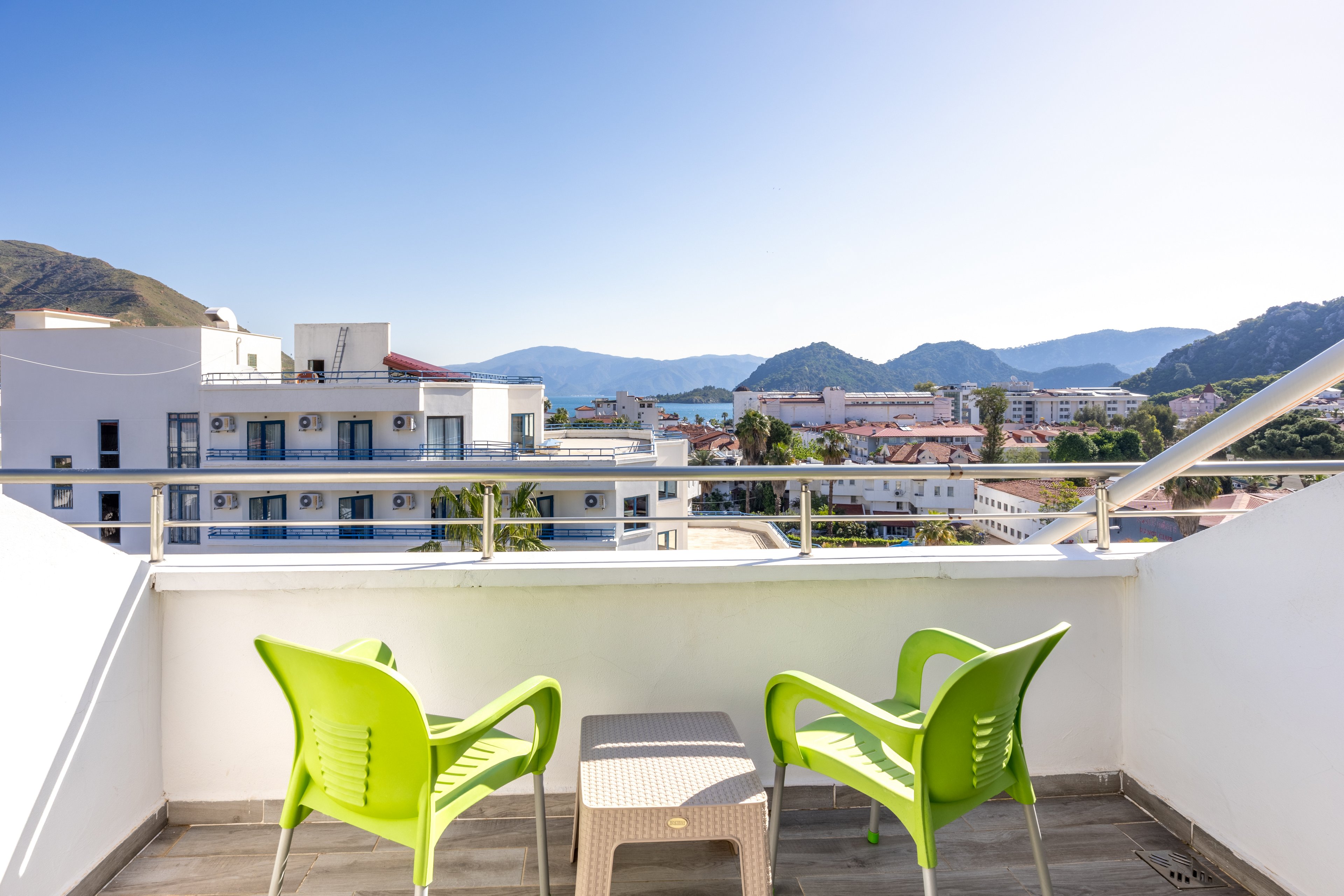 A bright balcony features two modern green chairs and a small table, overlooking a cityscape with distant mountains. The setting is outdoors, under clear blue skies, suggesting a warm and inviting atmosphere. White railings frame the view, and residential buildings are visible below. The overall mood is relaxed and tranquil, ideal for leisure or conversation.