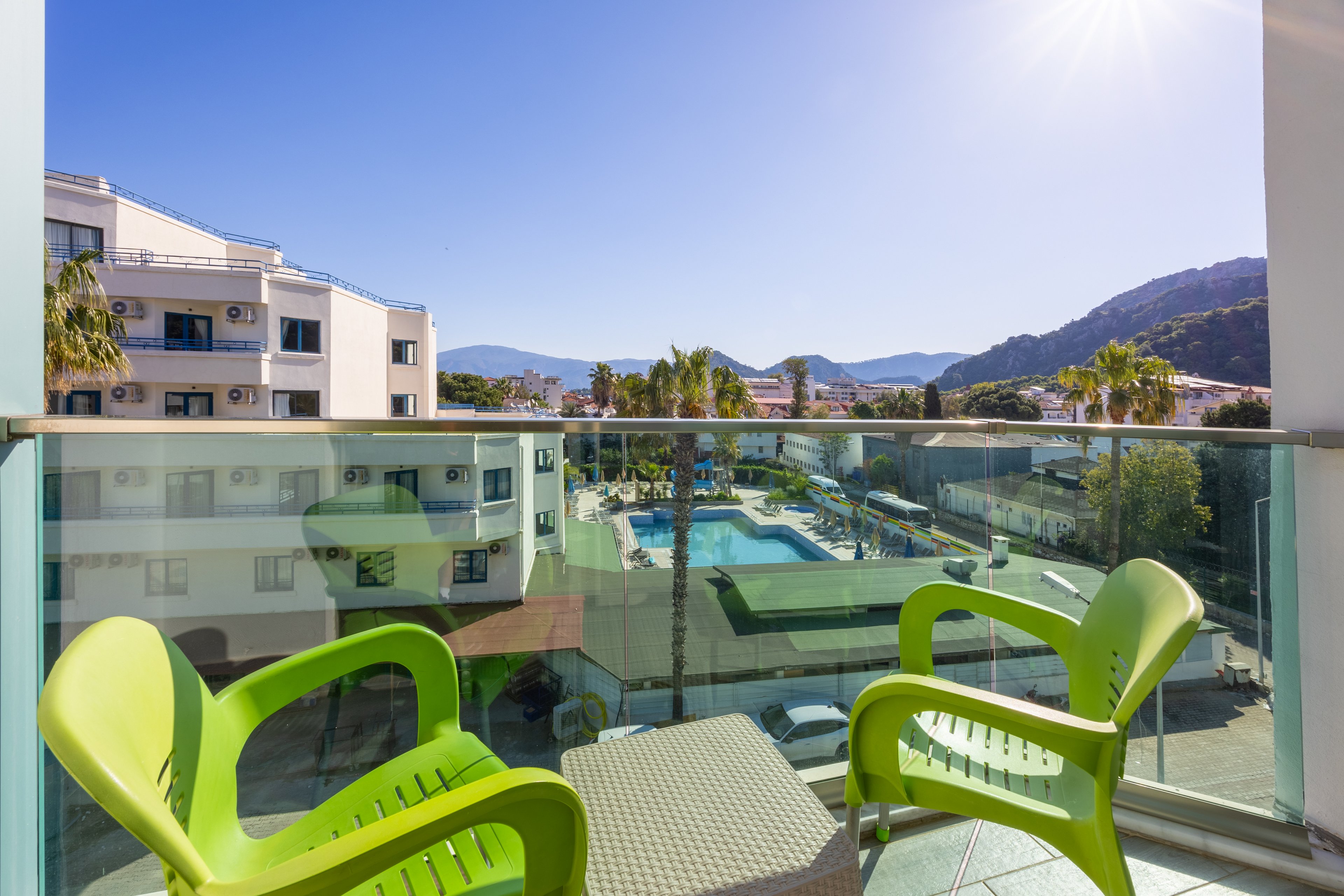A modern hotel balcony features two bright green chairs and a small table, overlooking a swimming pool and surrounding buildings. The scene is set on a clear, sunny day with blue skies and distant mountains. Palm trees and sunlit architecture create a relaxed, vacation atmosphere. The glass railing provides an unobstructed view of the outdoor pool area below. No visible text or numbers are present in the image.