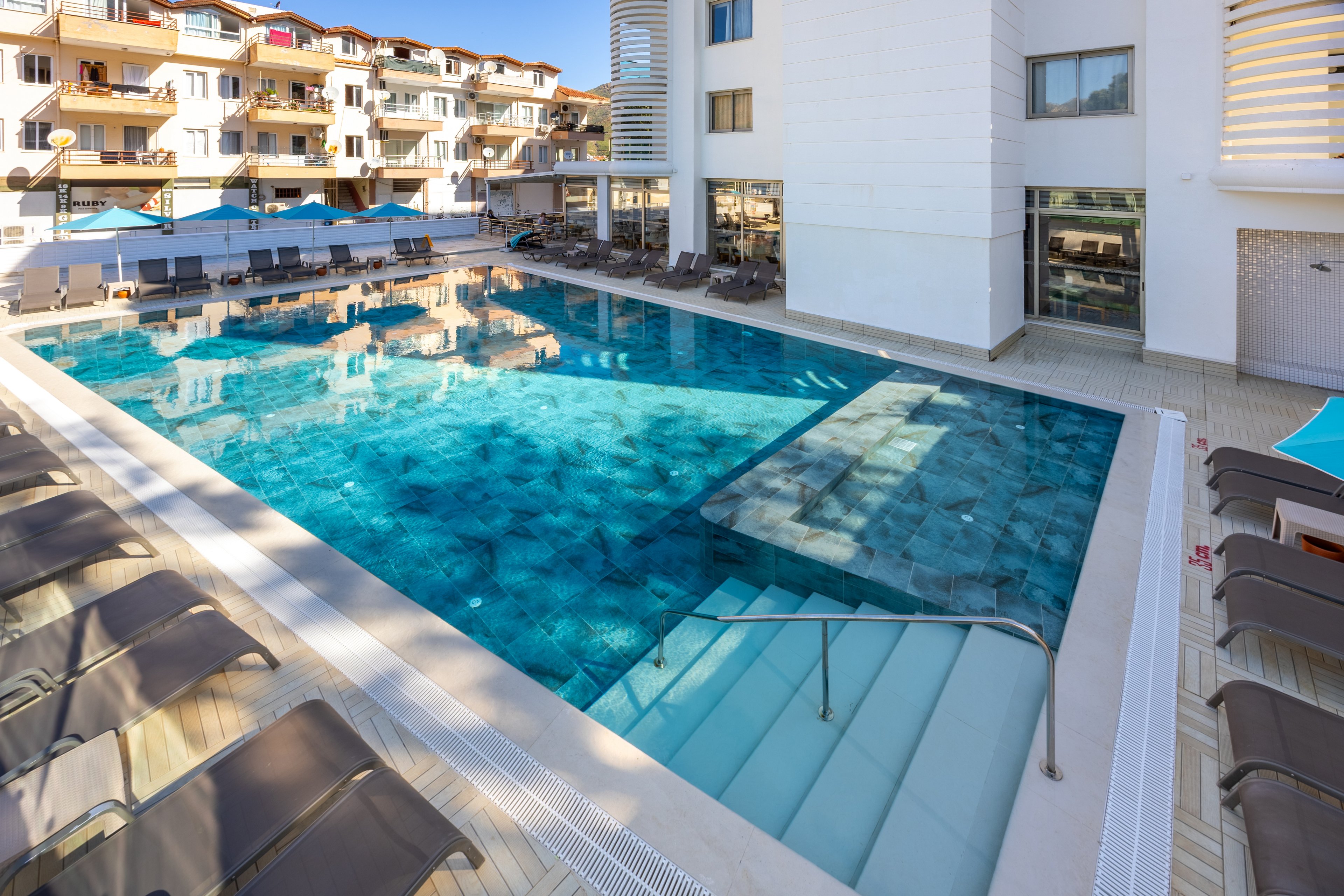 A large, clear swimming pool is the central feature of a modern hotel courtyard. Sun loungers line the poolside, and apartment-style balconies overlook the area. The setting is bright and sunny, with clean architectural lines and a relaxed, inviting atmosphere. No visible text or numbers are present in the image.