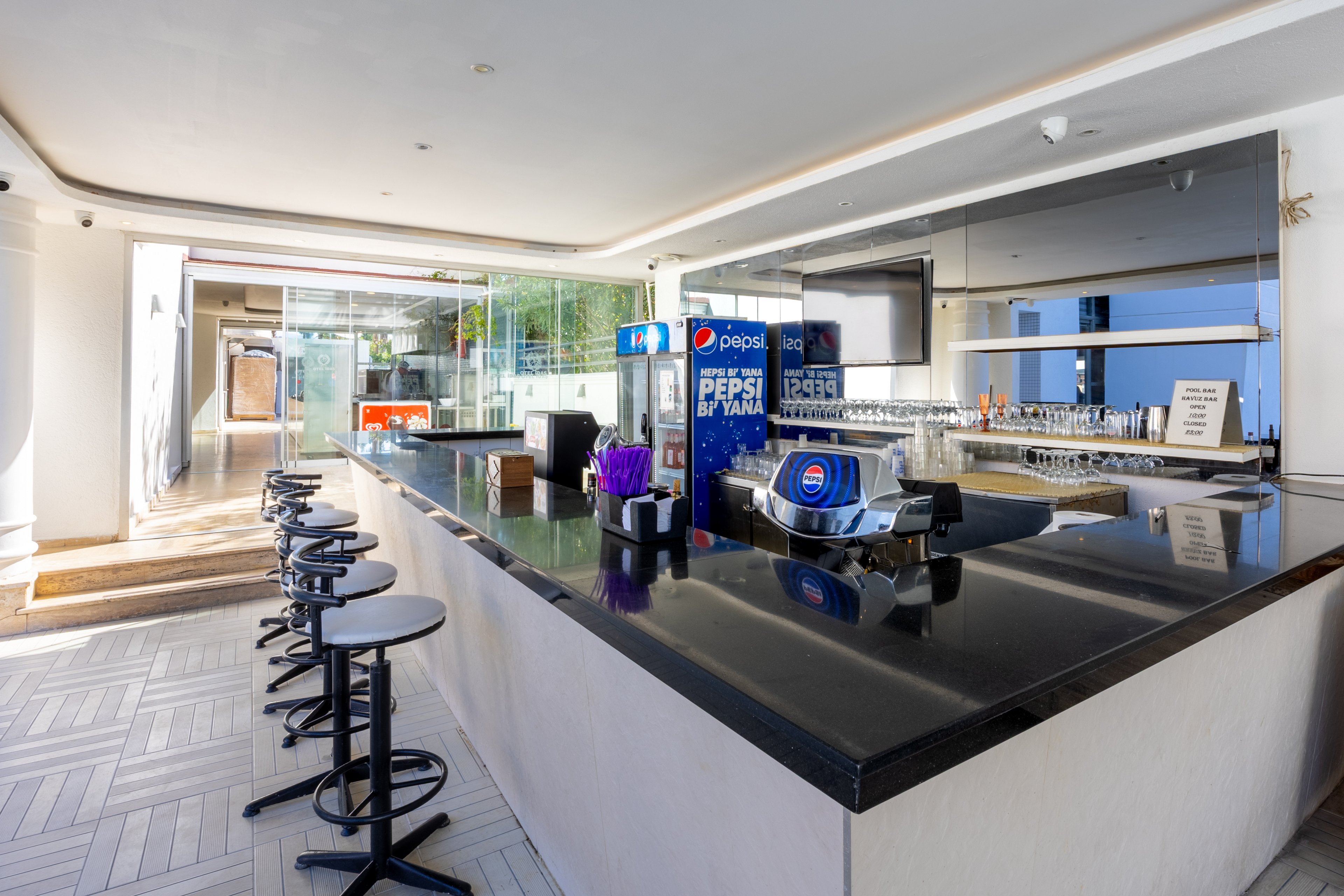 A sleek, contemporary hotel bar features a glossy black countertop and several bar stools lined up along the counter. A Pepsi soda dispenser and a napkin holder with the Pepsi logo are clearly visible on the counter, along with a numeric display showing '24:43'. The space is brightly lit with natural light streaming in from large windows, creating a clean and inviting atmosphere.