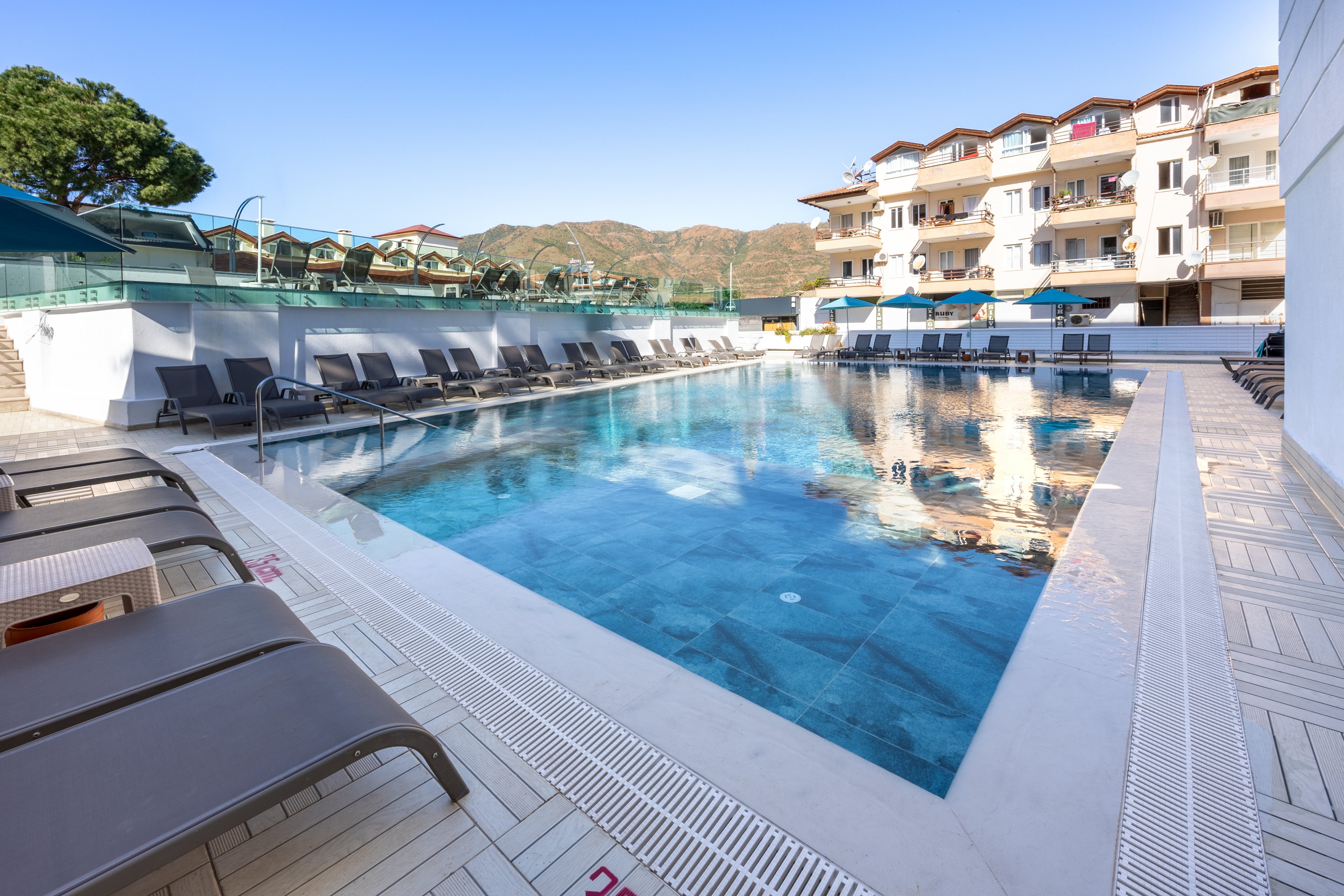 A clear, rectangular swimming pool is surrounded by modern lounge chairs at a hotel or resort. The setting is outdoors with mountains in the background and multi-story buildings reflecting in the water. The pool deck is clean and spacious, with visible numbers '120' and '220' marked on the edge. The atmosphere is calm and inviting, ideal for relaxation under a bright blue sky.