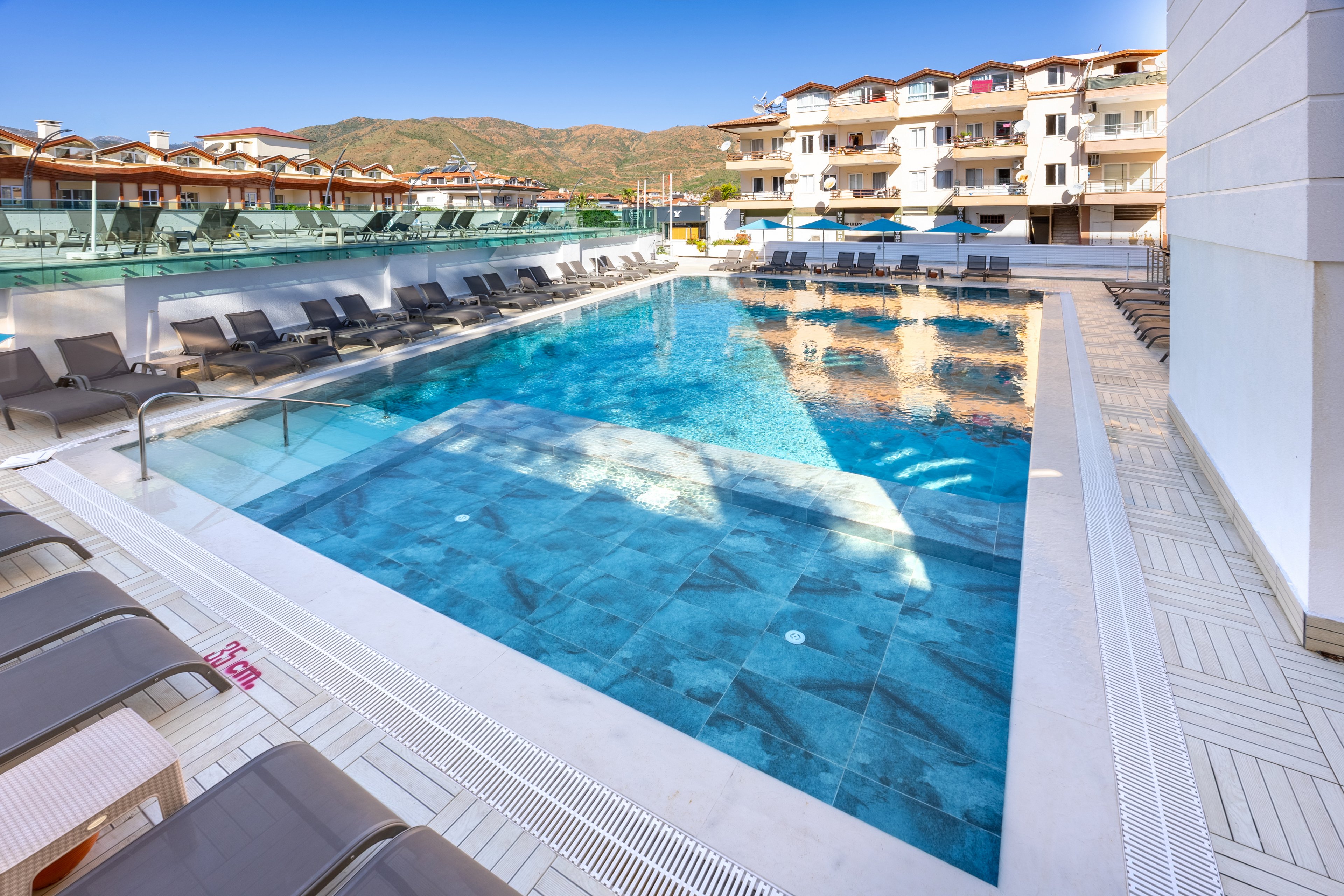 A contemporary outdoor swimming pool is surrounded by lounge chairs at a hotel or resort. The setting features clear blue water, tiled pool deck, and a scenic backdrop of mountains and multi-story buildings. The atmosphere is bright and inviting, with a clean, minimalist design and a calm, sunny mood. The pool area is empty, emphasizing relaxation and tranquility. The number '150 cm' is visible on the pool edge, indicating water depth.