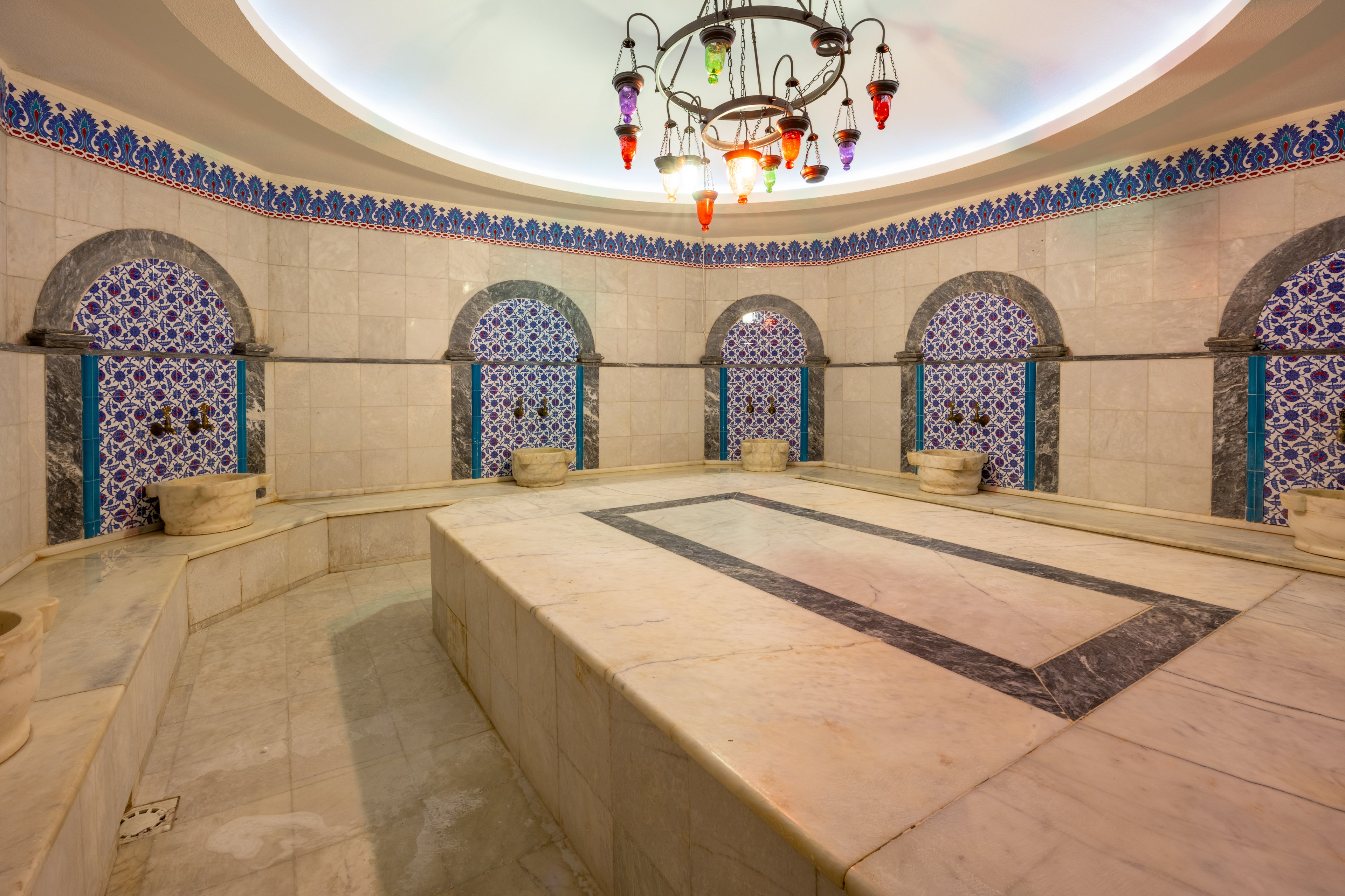 This image showcases the interior of a traditional Turkish hammam, featuring a large central marble platform surrounded by ornate tiled alcoves. The setting is illuminated by a decorative chandelier, highlighting the intricate blue and white tilework. The space is empty, emphasizing the serene and clean atmosphere typical of a bathhouse. The overall palette is neutral with pops of color from the tiles and lighting.