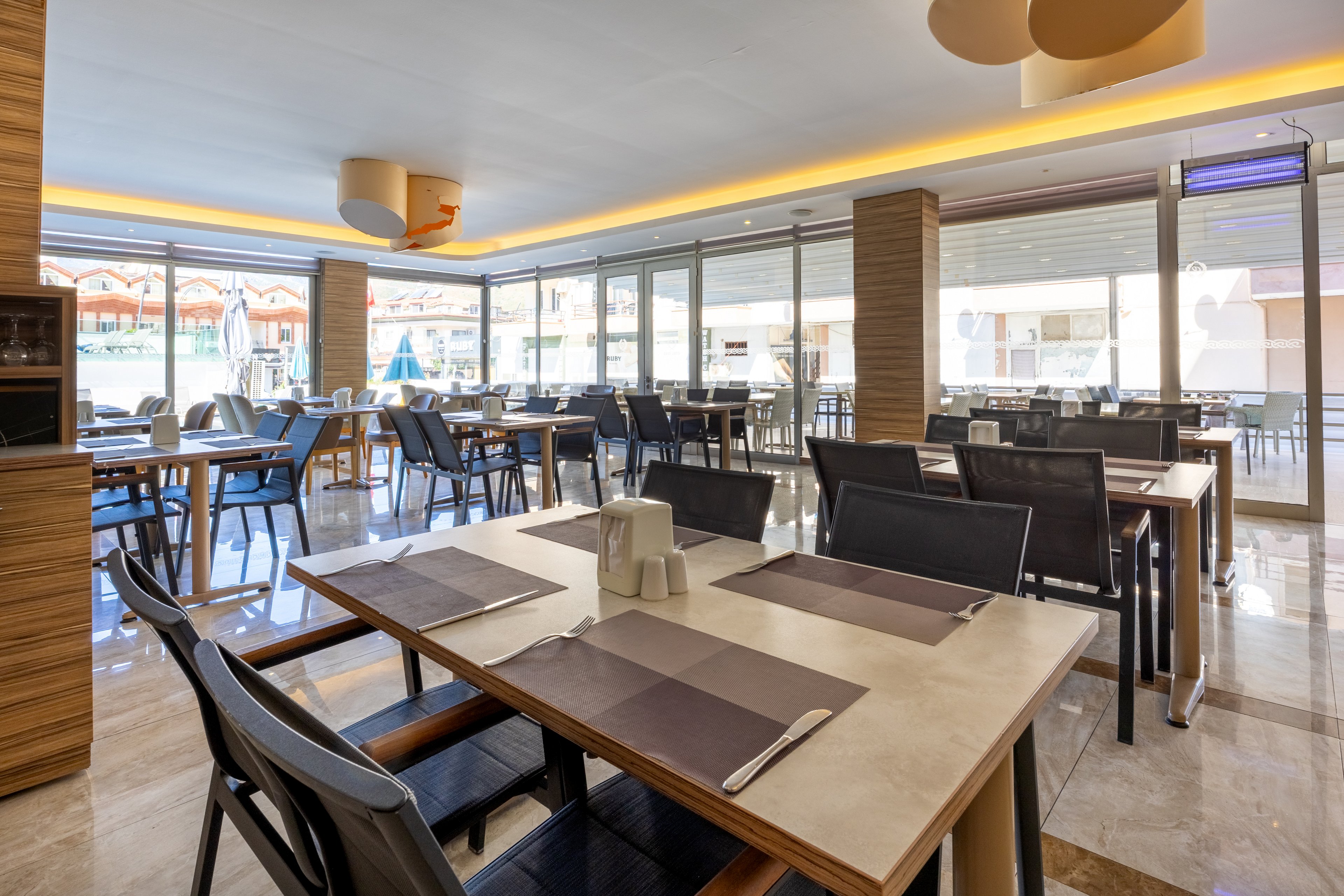 A spacious, contemporary restaurant dining area features neatly arranged tables and chairs, set with placemats and cutlery. Large windows allow natural sunlight to fill the room, creating a bright and inviting atmosphere. The interior design includes wood accents, neutral tones, and minimalist decor, contributing to a clean and modern look. No people are present, emphasizing the serene and orderly setting.