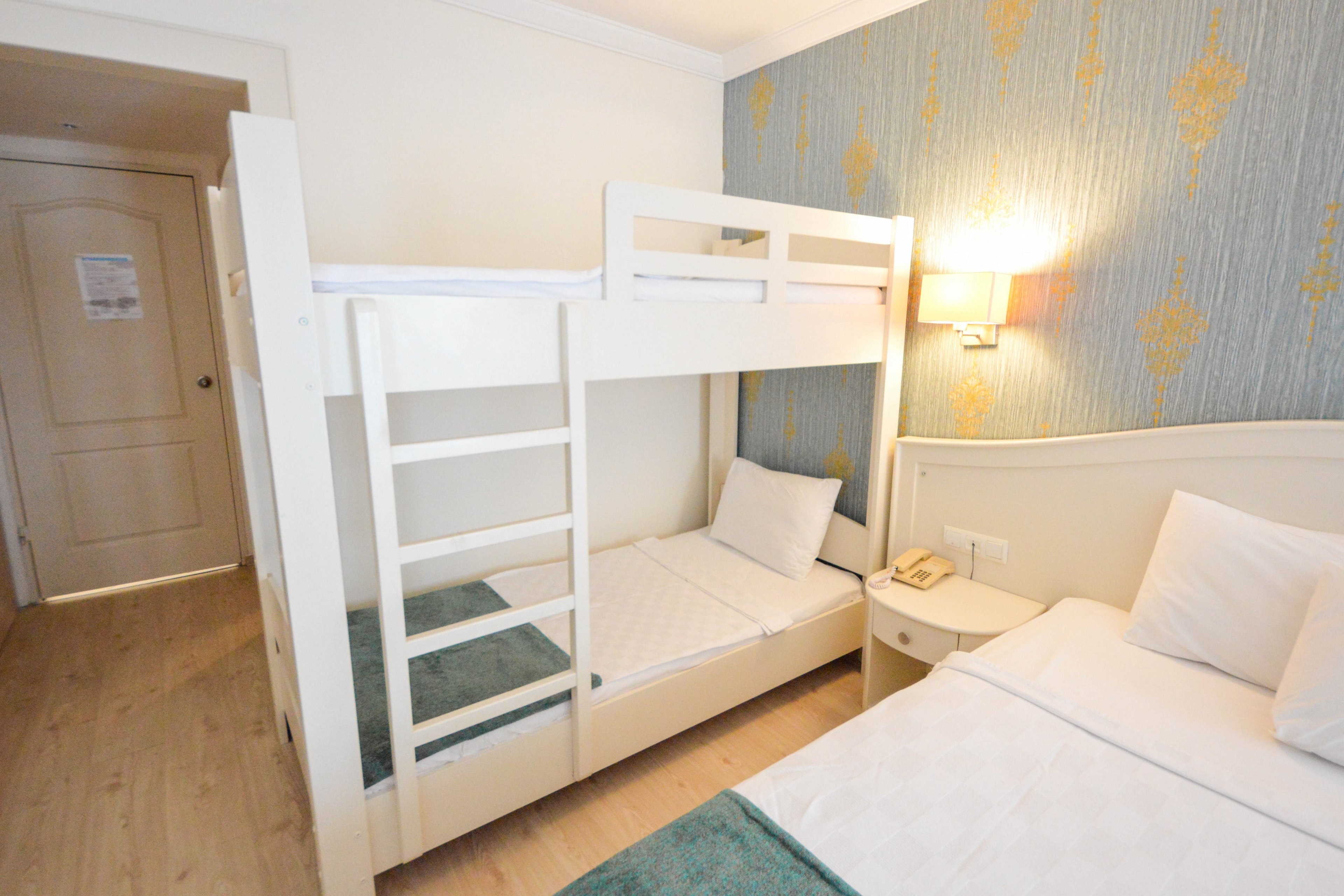 The image shows a bright hotel room featuring a white bunk bed and a single bed, both with crisp white linens. The setting includes a bedside table with a telephone and a wall-mounted lamp, against a blue accent wall with gold patterns. The room has light wood flooring and a door with a visible sign, creating a clean and welcoming atmosphere. No visible numeric values or brand names are present.