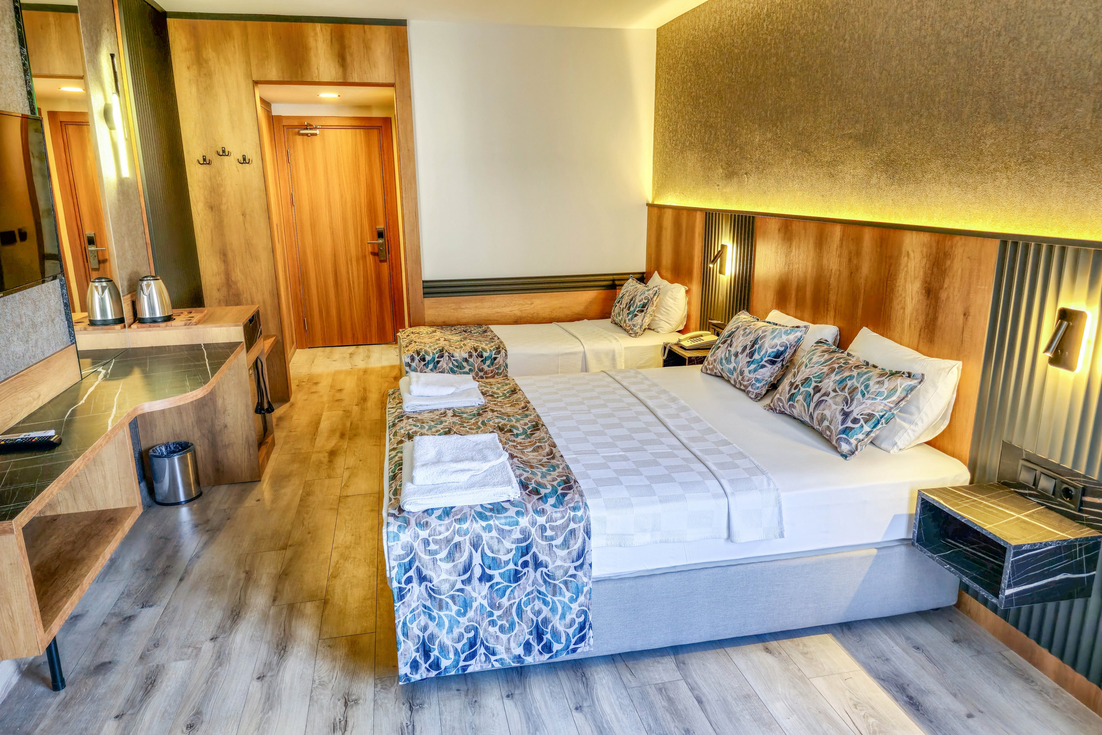 A contemporary hotel room features a double bed and a single bed, both adorned with patterned pillows and throws. The setting includes wood accents, a desk area with a kettle, and soft lighting that creates a welcoming atmosphere. The room is clean and organized, with towels neatly placed on the beds and a light wood floor. No visible text or numeric values are present in the image.