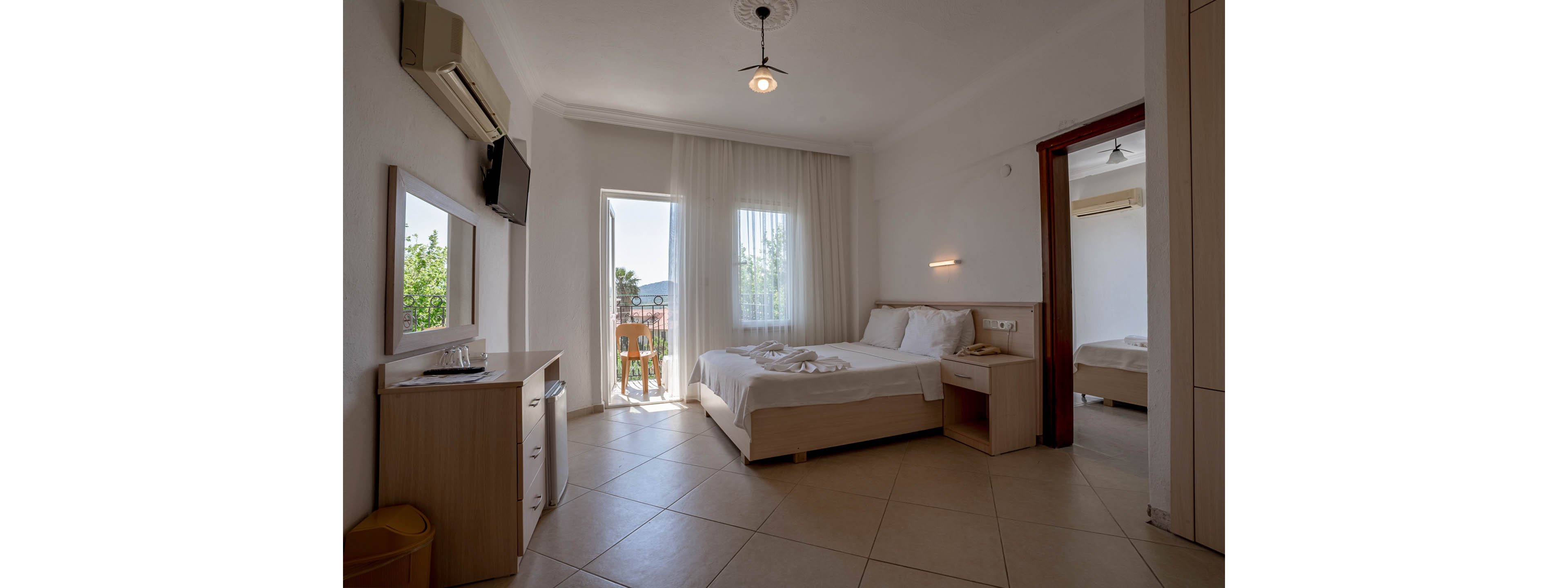 A tidy hotel bedroom features a double bed with white linens and towel arrangements at the center of the room. Natural light streams in through a glass door leading to a small balcony with a wooden railing and greenery outside. The interior includes a wall-mounted television, air conditioner, desk with mirror, and neutral-colored tile flooring. An adjacent room with another bed is visible through an open sliding door, enhancing the sense of space and comfort.