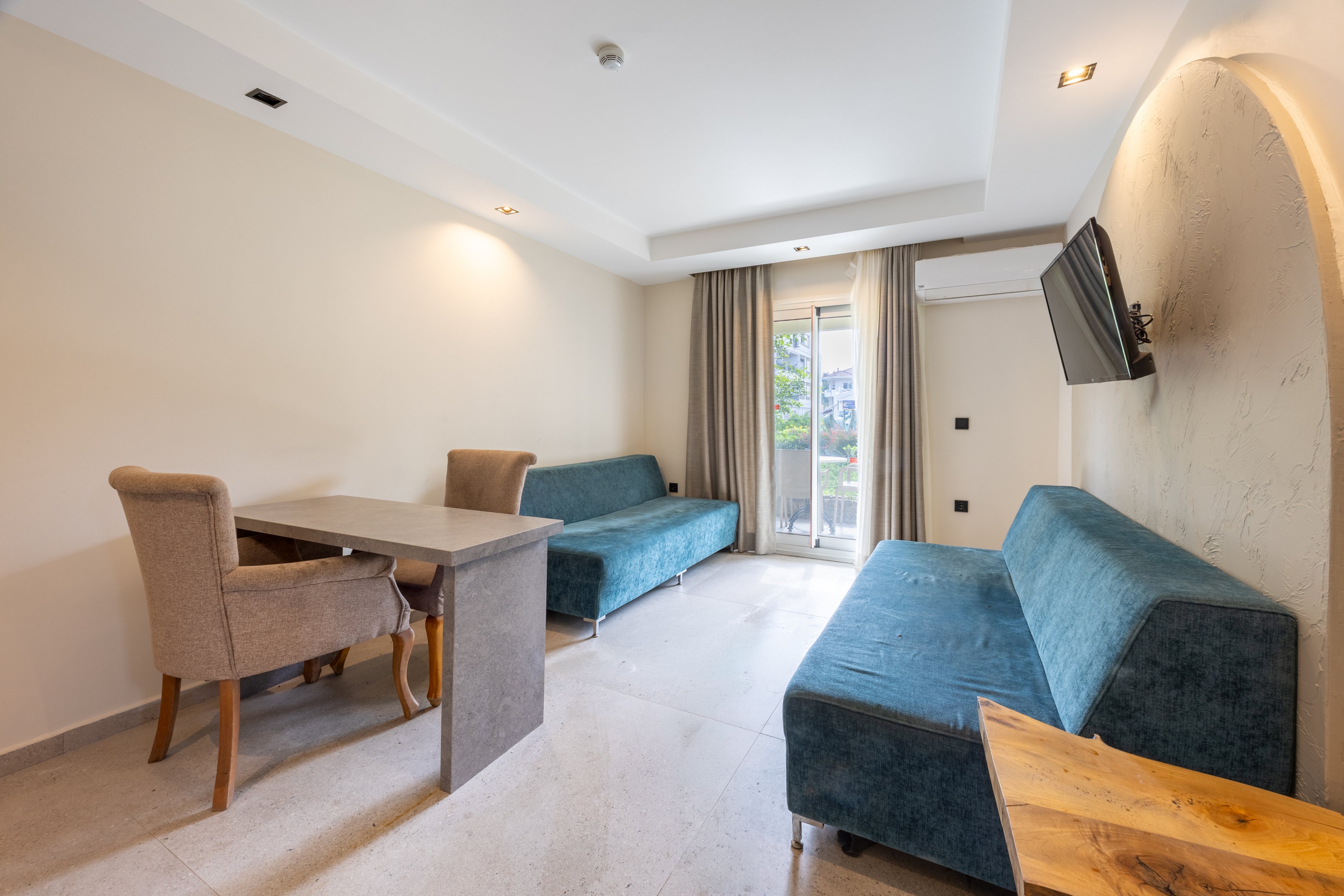A contemporary hotel room features two teal sofas, a small dining table with upholstered chairs, and a wall-mounted television. The space is well-lit with natural light coming from a glass door leading to a balcony with outdoor greenery visible. Neutral tones dominate the palette, creating a calm and inviting atmosphere, while the room appears tidy and uncluttered.
