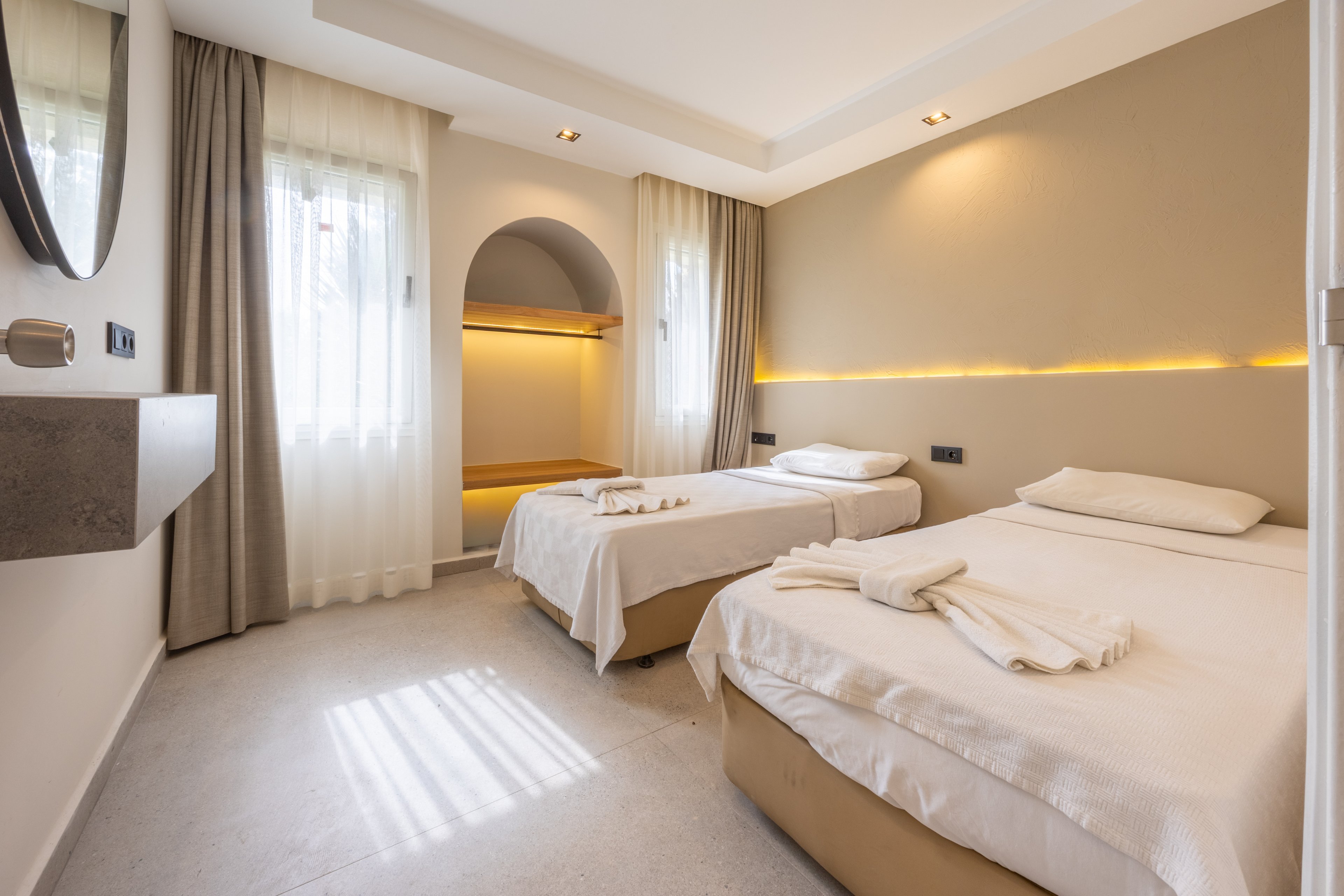 The image shows a contemporary hotel room featuring two single beds with crisp white linens and neatly folded towels. The setting is bright, with natural light streaming through sheer curtains and warm accent lighting on the wall. The room includes minimalist decor, neutral tones, and a small alcove with shelving. No visible text or numbers are present in the image.