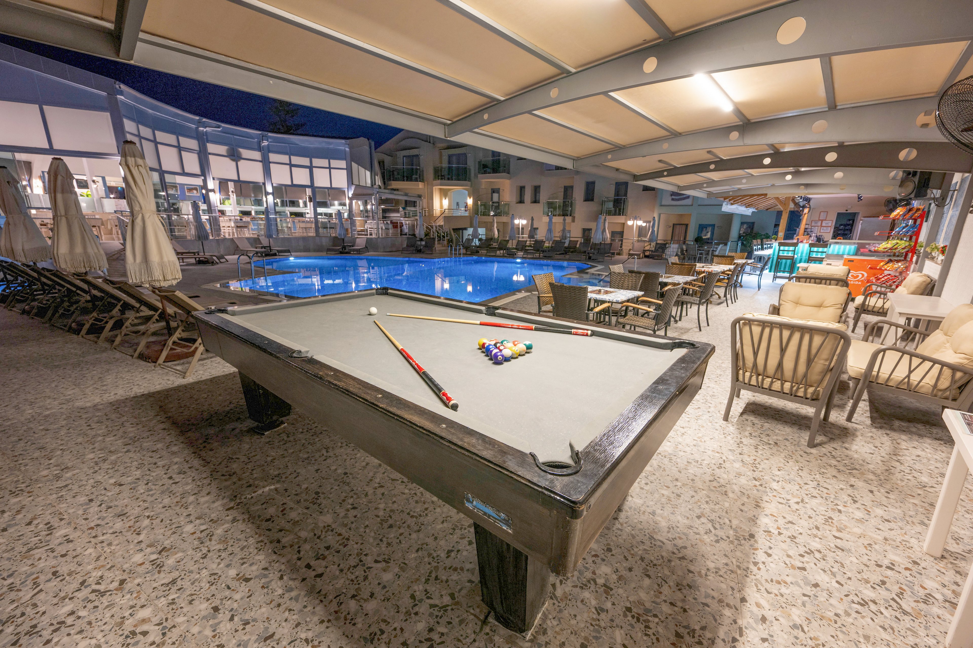 An outdoor billiards table with balls racked and a cue stick rests under a covered terrace. The scene overlooks a brightly lit swimming pool surrounded by lounge chairs and umbrellas. Nearby, tables and cushioned chairs create a relaxed seating area for guests. The image is taken at night, giving the resort setting a calm and inviting atmosphere.