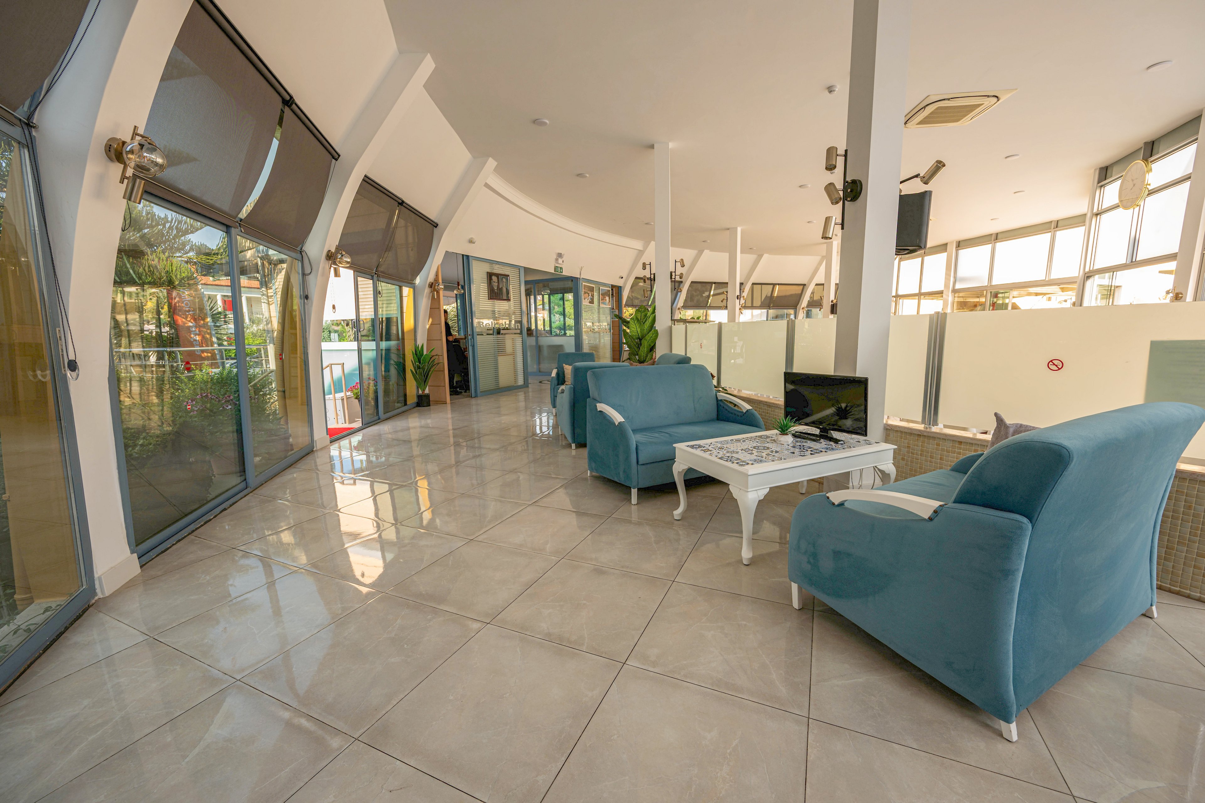 A spacious modern lounge features blue upholstered armchairs arranged around a small white coffee table with a television on top. Large floor-to-ceiling windows line one side of the room, allowing abundant natural light to flood the tiled floor. Potted plants and glass partitions add a touch of greenery and separation within the open-plan interior. The overall atmosphere is bright, airy, and contemporary, suitable for a waiting area or communal relaxation space.