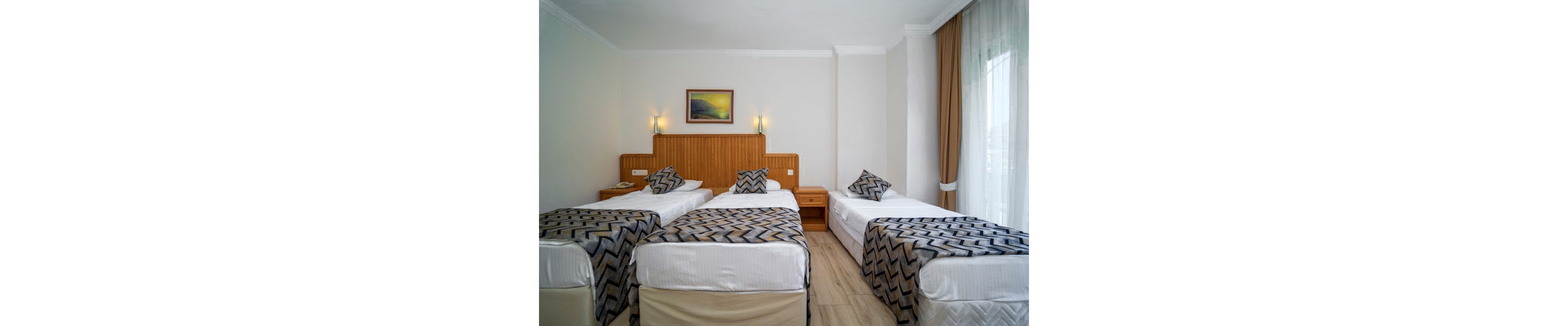 A tidy hotel room features three single beds arranged side by side. Each bed has white linens and patterned gray and black throws with matching cushions. The room includes a wooden headboard, wall-mounted lights, and a framed landscape picture above the beds. Natural light enters through large windows with sheer curtains and beige drapes, highlighting the clean, modern interior.