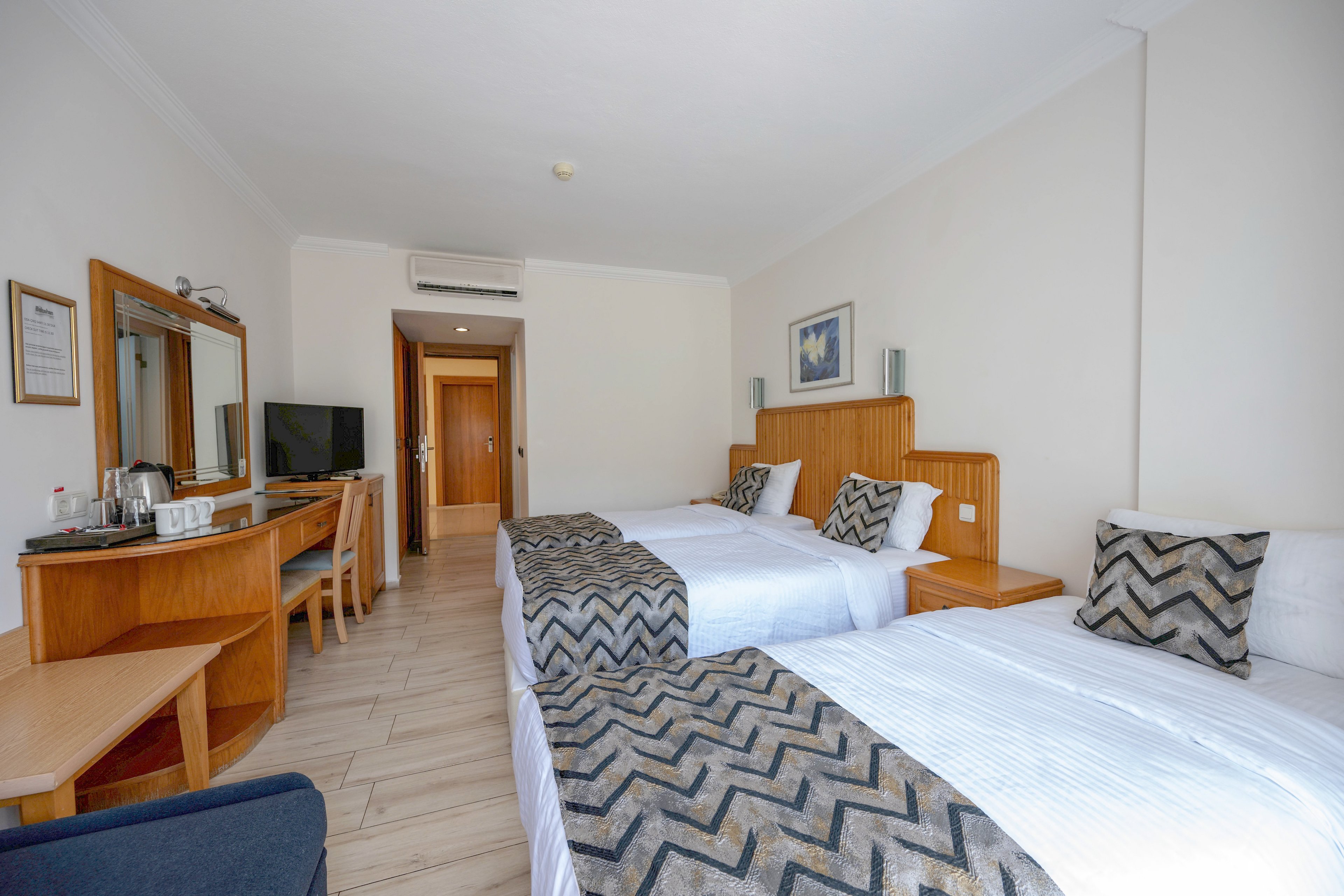 A modern hotel room features two neatly made twin beds with patterned throws and matching cushions. The space includes a wooden desk and vanity area with a flat-screen TV, mirror, and tea and coffee set. Light-colored walls and flooring create an airy, spacious feel, complemented by natural light from an unseen window. A doorway at the back leads to the corridor, emphasizing the room’s clean and organized layout.