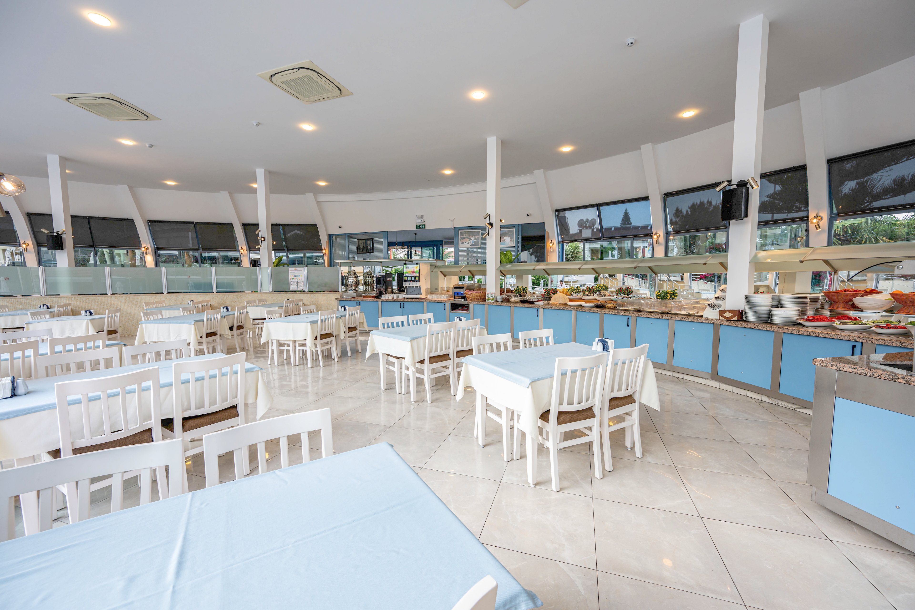 Spacious self-service restaurant interior with neatly arranged white tables and chairs. A long buffet counter lines one side of the room, displaying various dishes, plates, and serving equipment. Large windows and ceiling lights create a bright, airy atmosphere with clean blue and white tones. The dining area appears empty and ready for guests, emphasizing order and freshness.