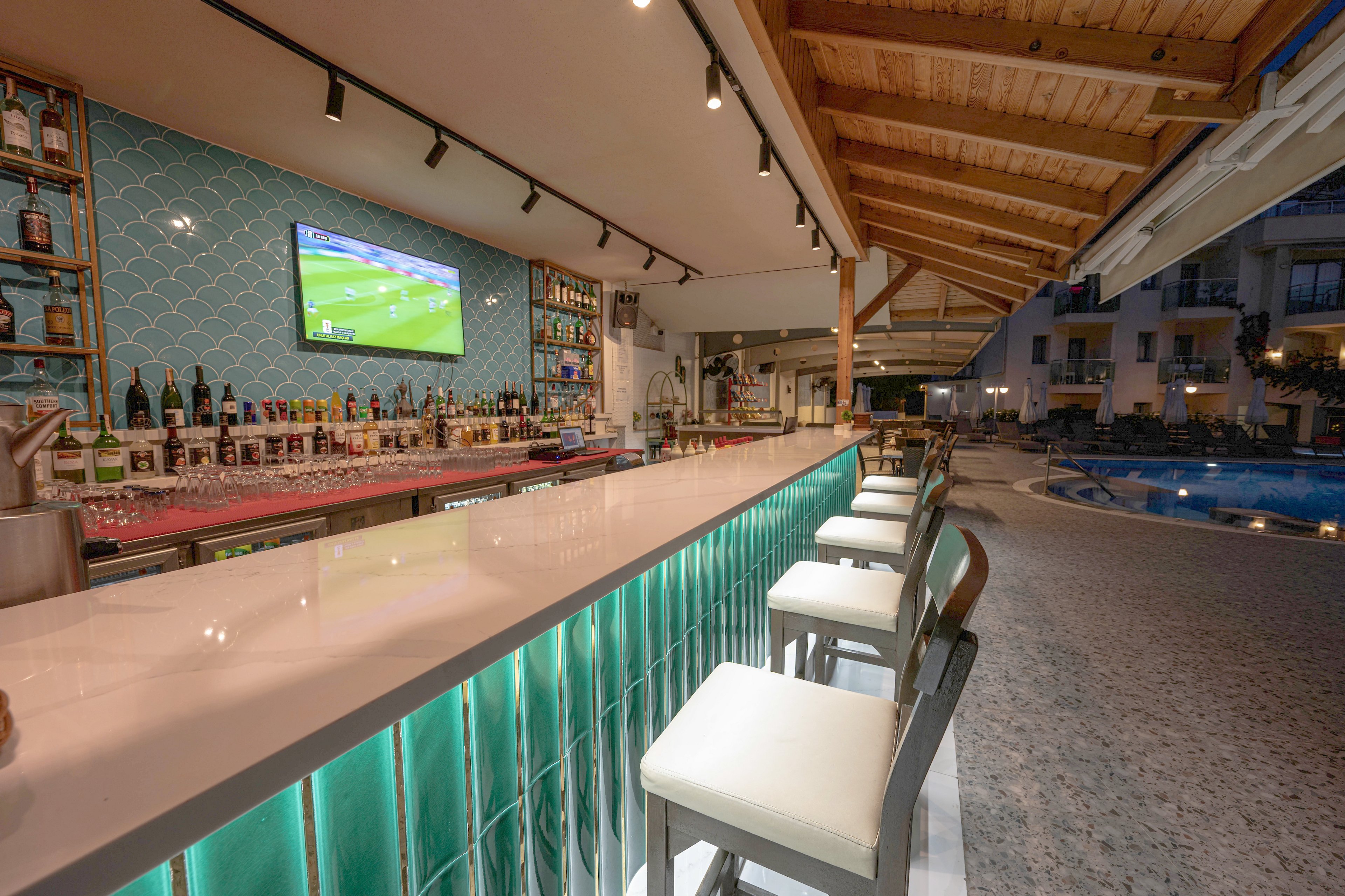 A sleek, modern bar with a long white countertop is shown beside an outdoor swimming pool at dusk. High stools line the bar, which features glowing turquoise panels and a tiled backsplash. Bottles and glassware are neatly arranged on shelves, and a large television displays a soccer match above the bar. The setting appears relaxed and inviting, with warm lighting and resort buildings visible in the background.