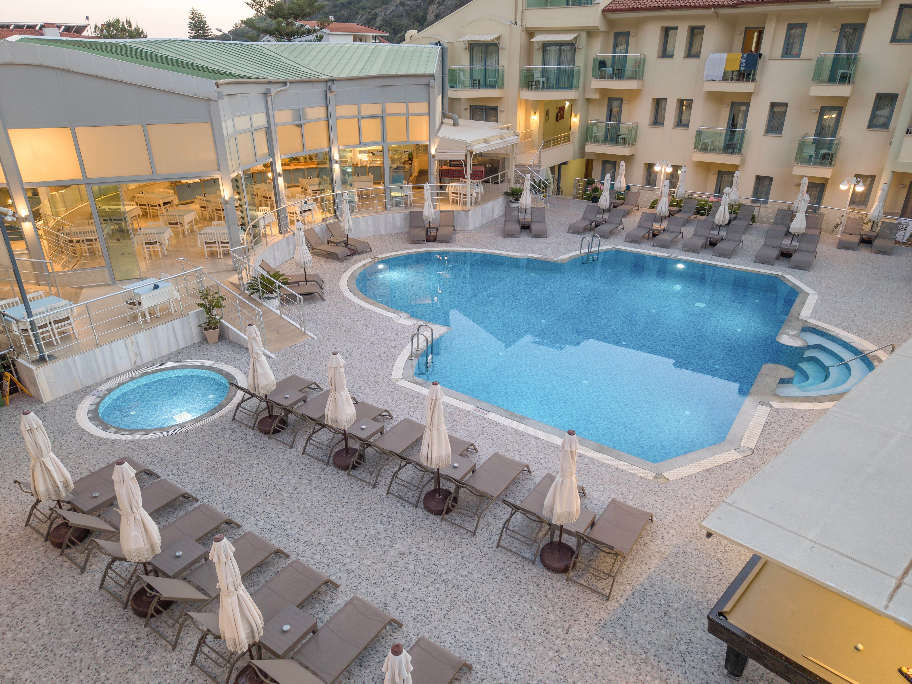An elevated view shows a modern hotel courtyard centered around a large, uniquely shaped swimming pool. Numerous sun loungers and closed umbrellas surround the pool, with an additional small circular pool nearby. The adjacent building features multiple balconies and large windows, suggesting guest rooms overlooking the water. Soft evening lighting and calm, empty seating areas create a tranquil, resort-like atmosphere.