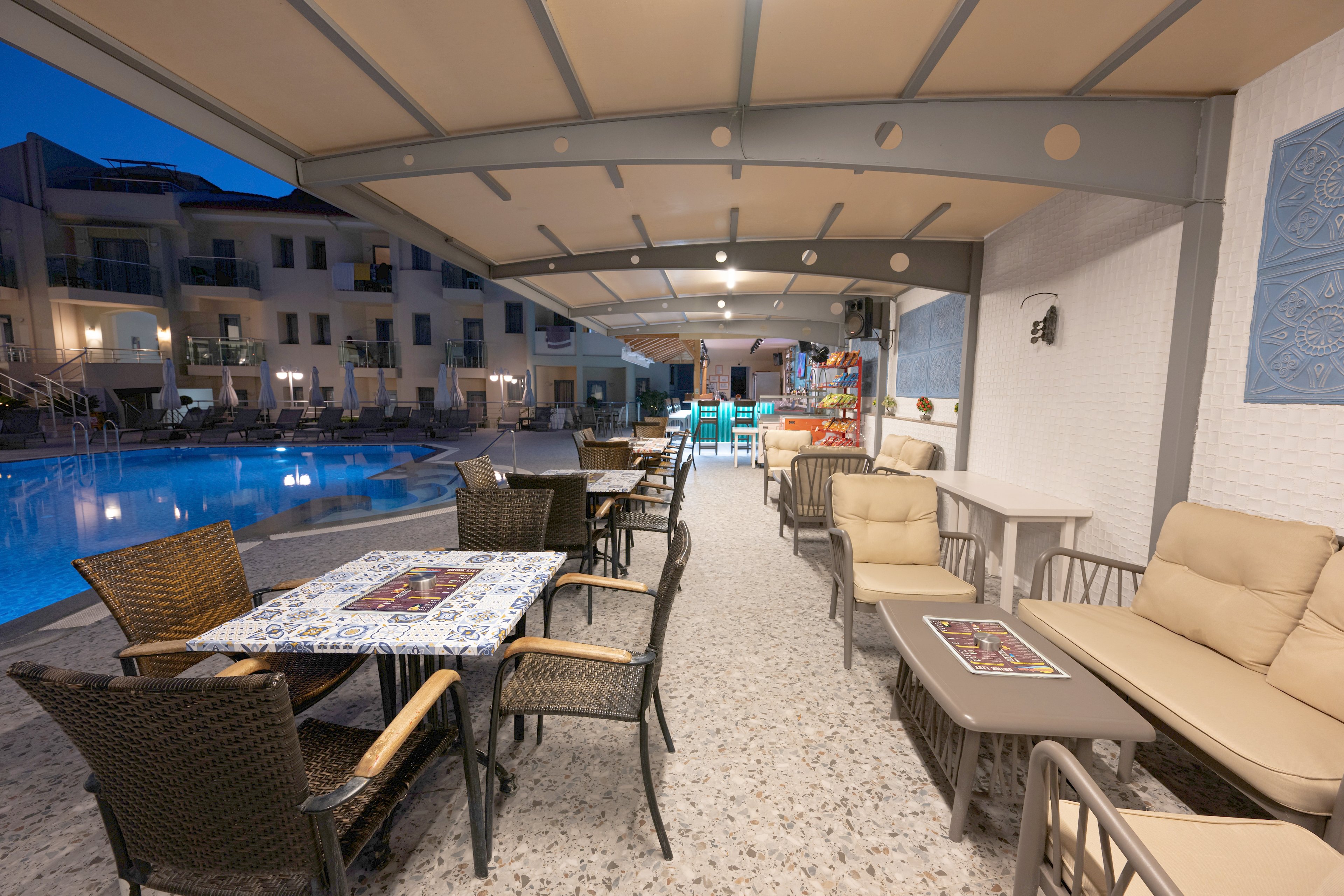 An inviting outdoor lounge and dining area is set beside a swimming pool in the evening. Wicker chairs and cushioned sofas surround tables with decorative tile-style surfaces under a covered terrace. Soft lighting reflects off the calm pool water, creating a relaxed resort atmosphere. Apartment-style buildings and additional seating are visible in the background, suggesting a hotel or vacation property.