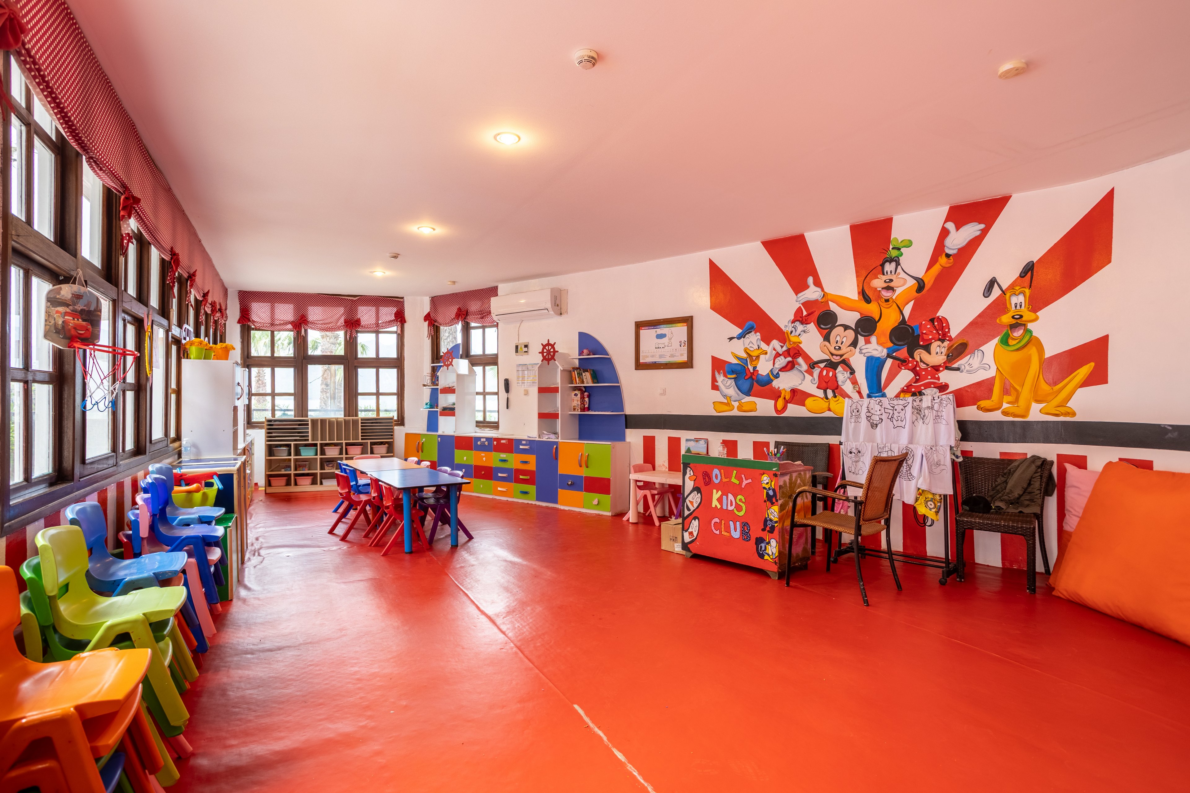 Brightly colored children's playroom with small plastic chairs and tables arranged along the walls. The room features a bold red floor, large windows with red curtains, and shelves filled with toys and books. A vibrant wall mural of cartoon characters decorates the far wall above a small activity area. The space appears designed for young kids’ activities, crafts, and group play in an indoor kids club setting.