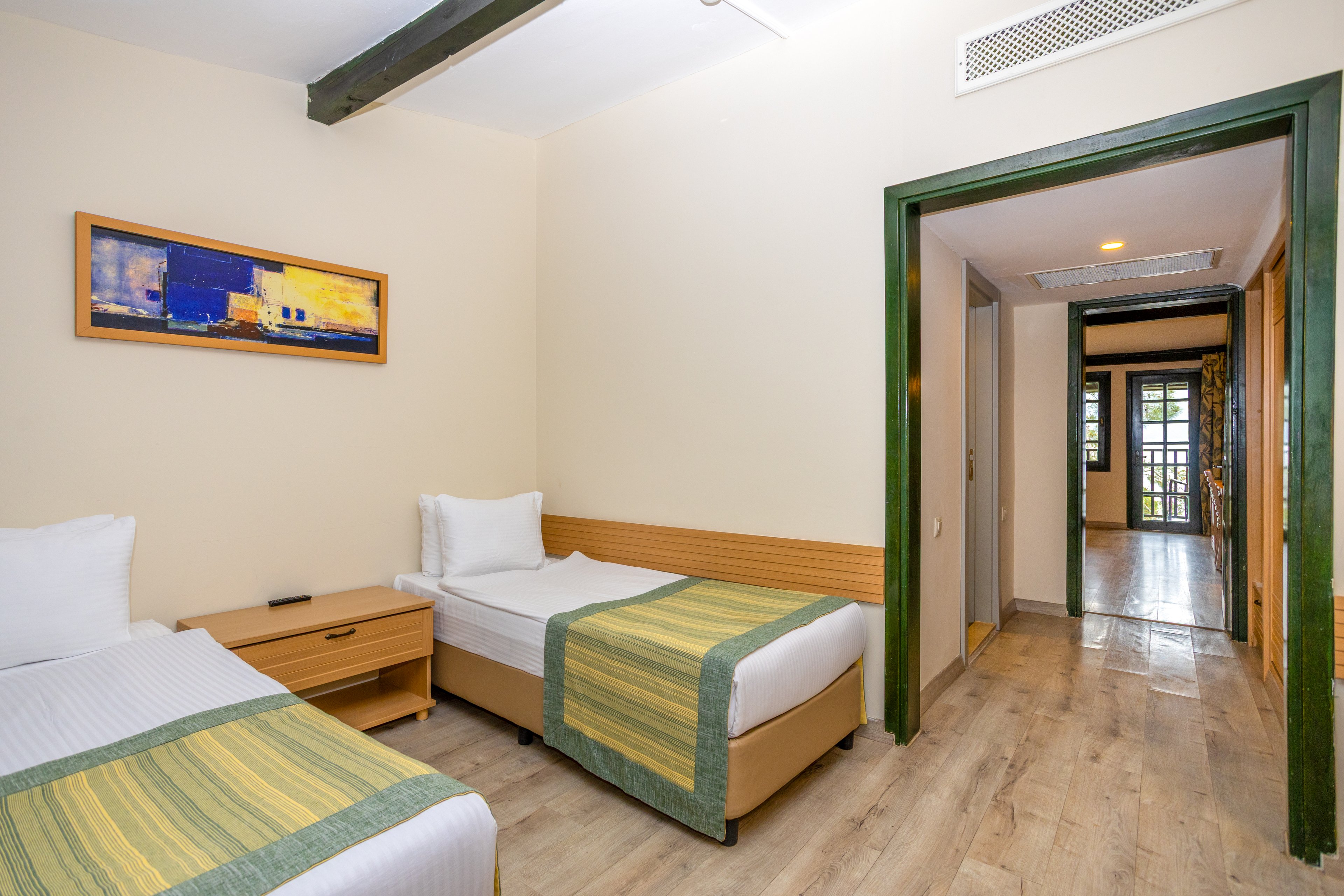 A bright hotel room features two single beds with white linens and green striped runners. A wooden nightstand sits between the beds, and a colorful abstract painting hangs on the wall above. The room opens into a hallway with wooden flooring leading to a doorway and windows at the far end. Soft lighting and neutral walls create a clean, welcoming atmosphere suitable for guests.