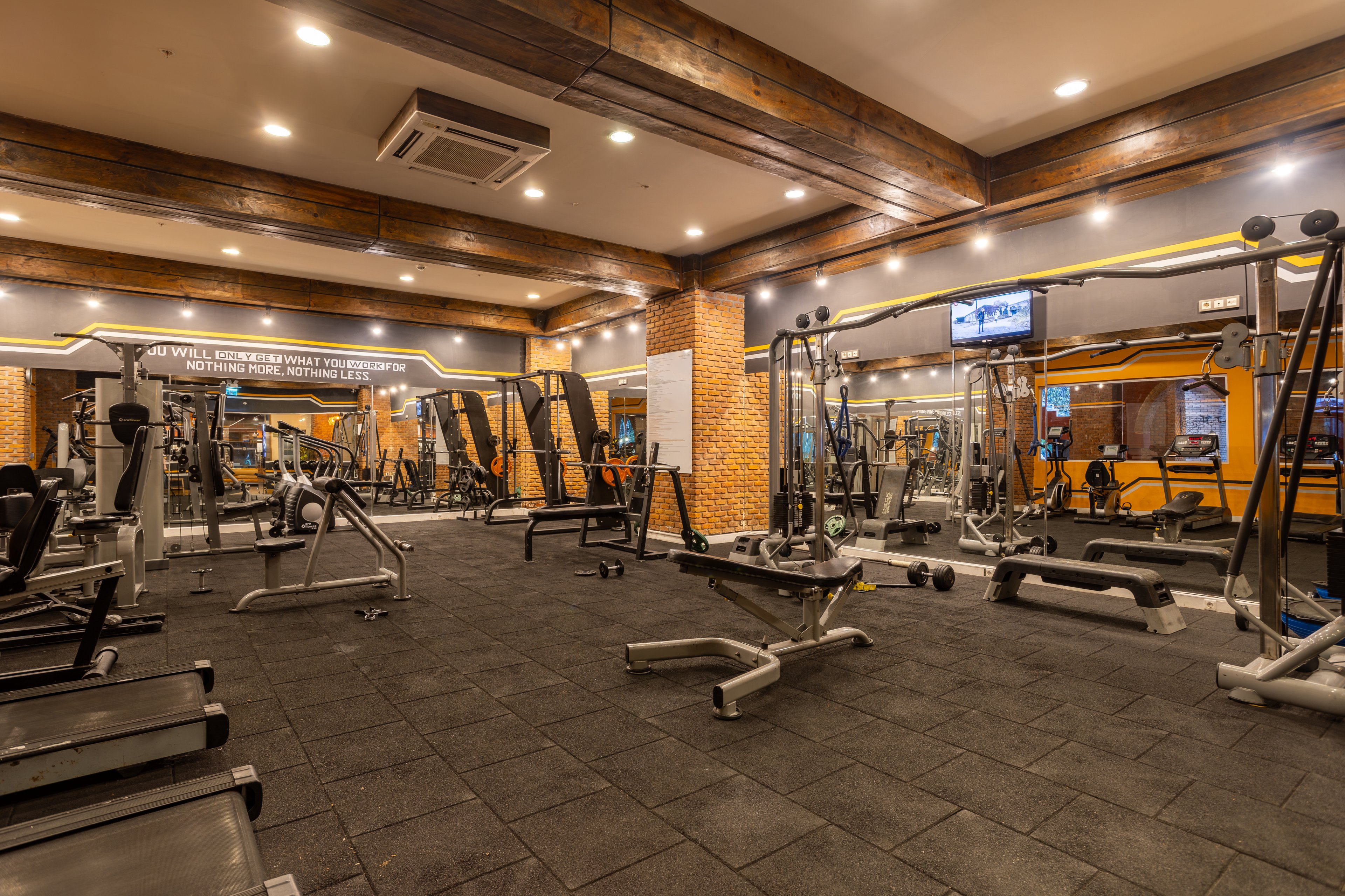 Spacious modern gym interior featuring various strength and cardio machines arranged on a rubberized floor. The room is empty, with no people present, emphasizing the equipment and clean workout environment. Warm ceiling lights, wooden beams, and brick columns create a cozy yet professional atmosphere, with large mirrors visually expanding the space. A wall-mounted television and bright yellow accent walls add contemporary details to the fitness center.