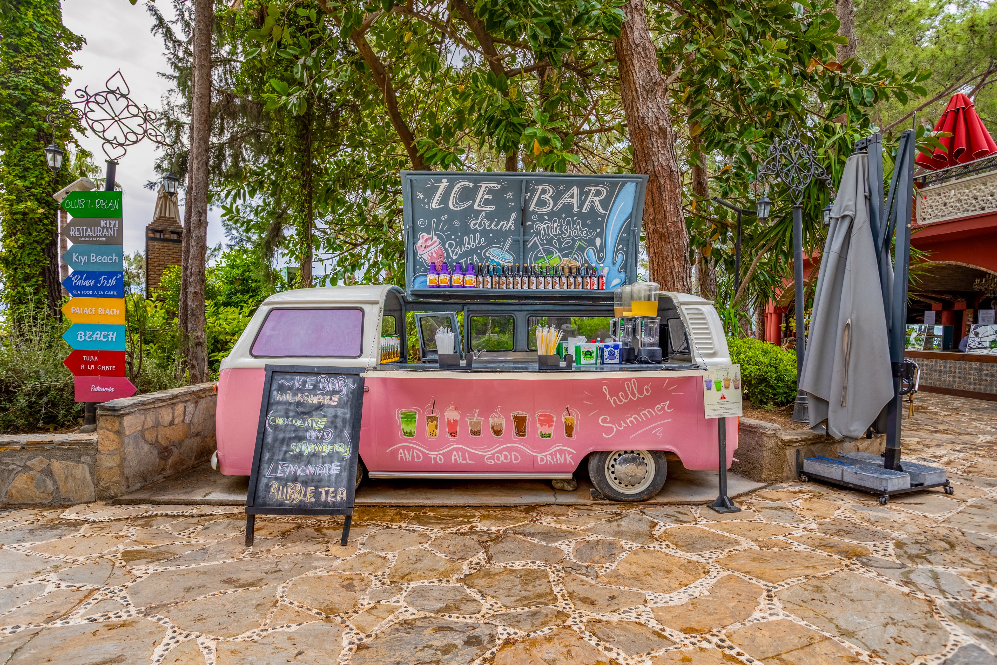A retro van converted into an ice bar serves colorful drinks in an outdoor setting. The pink vehicle is decorated with illustrations of beverages and the phrase "hello Summer" on its side. A chalkboard menu advertises bubble tea and other refreshments, while bottles and straws line the counter. Surrounding trees, stone pavement, and a multicolored direction sign create a relaxed vacation atmosphere.