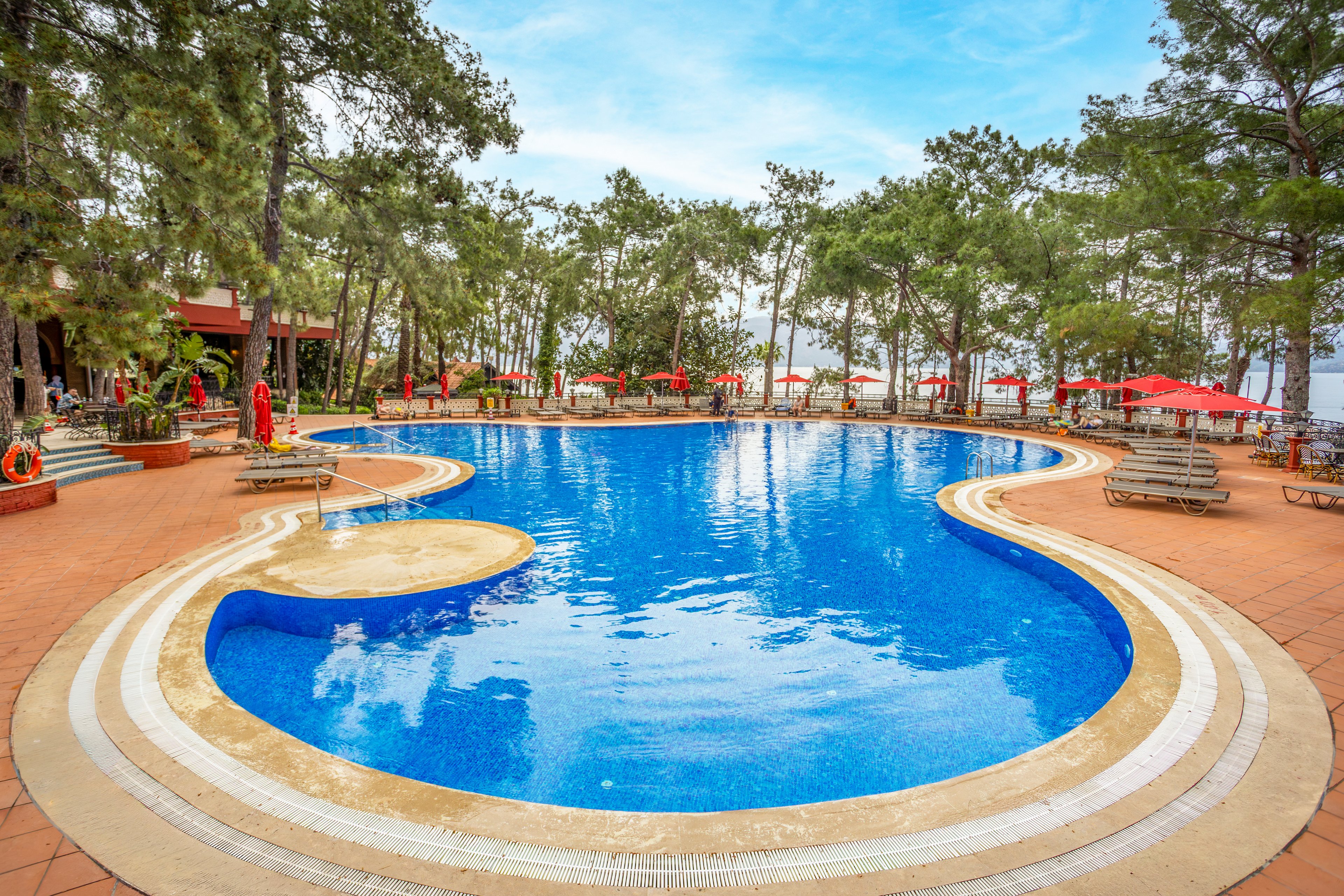 A large curved swimming pool is set outdoors within a wooded resort area. Tall trees surround the pool, creating a natural, shaded environment. Sun loungers and red umbrellas line the poolside terrace, suggesting a relaxing vacation setting. The bright blue water contrasts with the warm tiled deck under a partly cloudy sky.