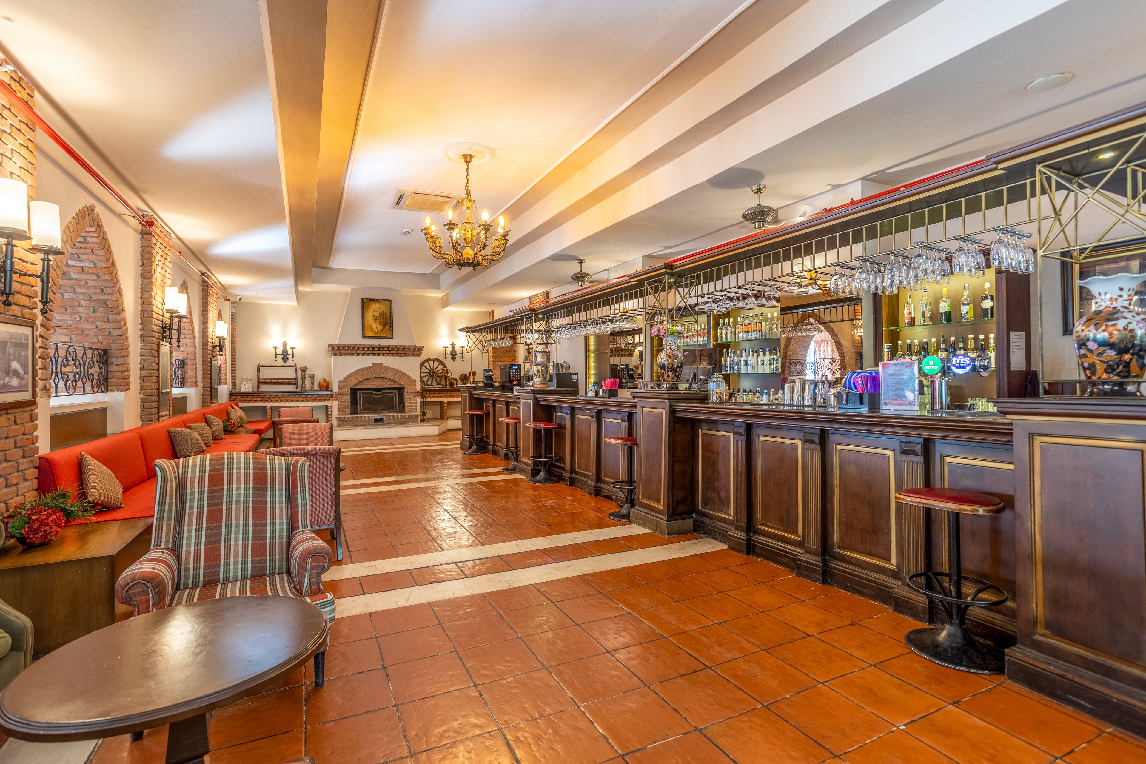 Spacious hotel bar interior featuring a long wooden counter and fully stocked shelves of bottles and glassware. The setting includes tiled floors, exposed brick walls, and a decorative fireplace at the far end of the room. Comfortable seating with upholstered armchairs and red cushioned benches lines the side, creating an inviting lounge area. Warm lighting from chandeliers and wall sconces enhances the cozy, traditional atmosphere.