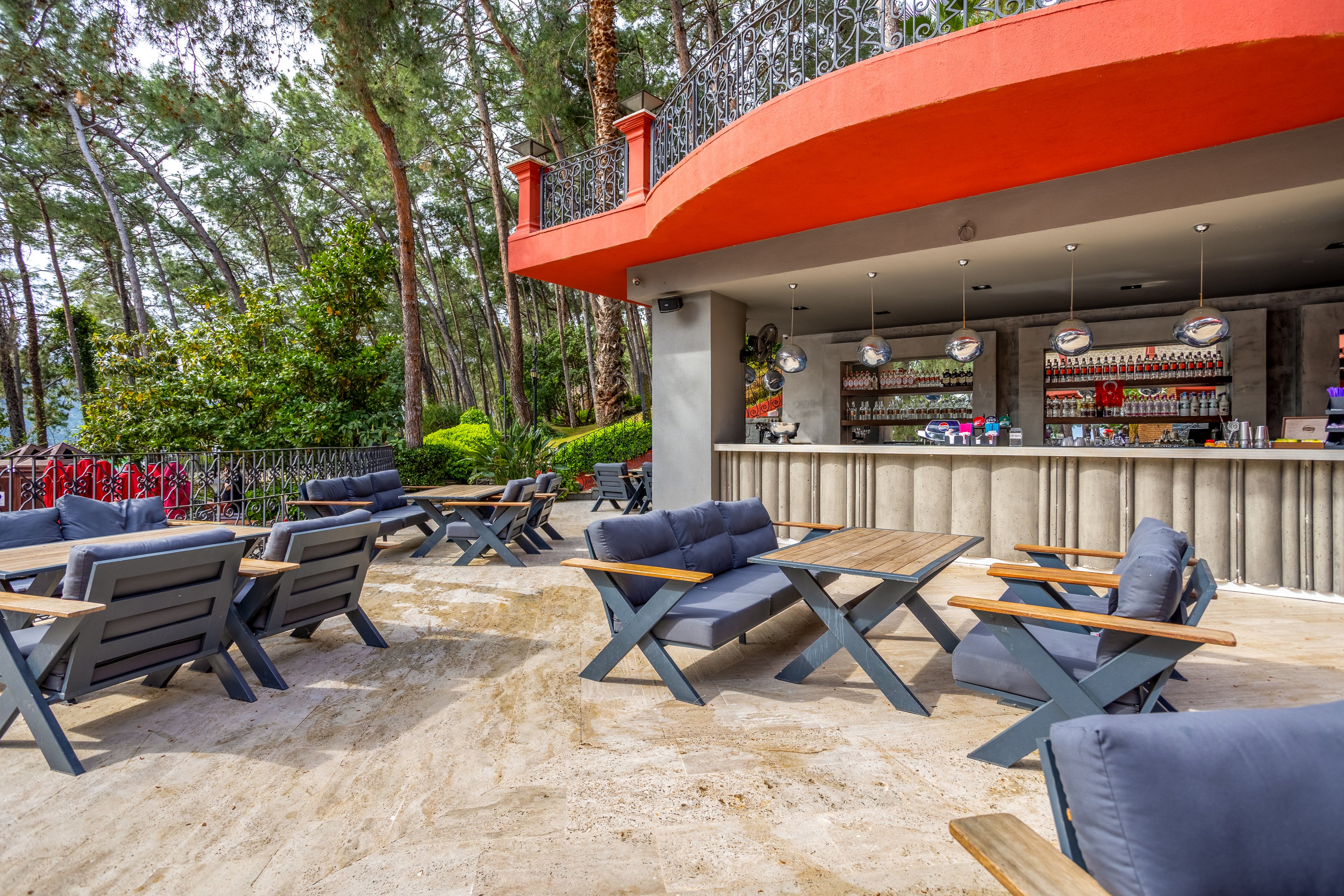 An open-air bar terrace is set among tall trees, featuring modern wooden tables and cushioned lounge chairs. The bar counter in the background is stocked with bottles and glassware, with hanging spherical lights above. The seating area is spacious and arranged for relaxed socializing on a stone or concrete patio. Lush greenery and a decorative railing frame the scene, creating a tranquil, resort-like atmosphere.