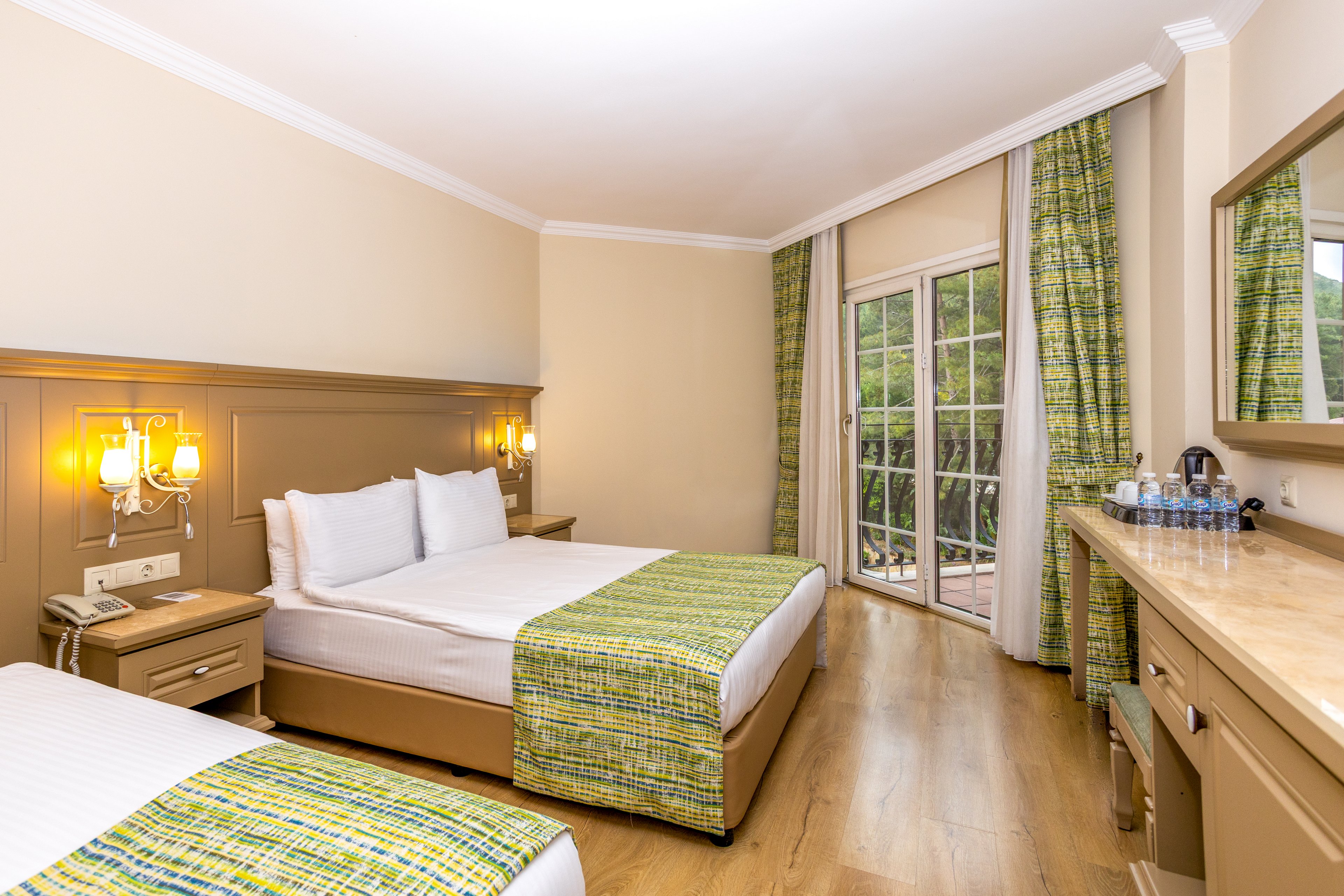 A clean, modern hotel room features two neatly made beds with white linens and green patterned throws. Warm wall sconces illuminate the headboard area beside a telephone and bedside controls. A large glass door opens to a balcony with a view of greenery outside, bringing in natural light. A long wooden desk with a mirror, chair, and bottled water completes the comfortable guestroom setting.