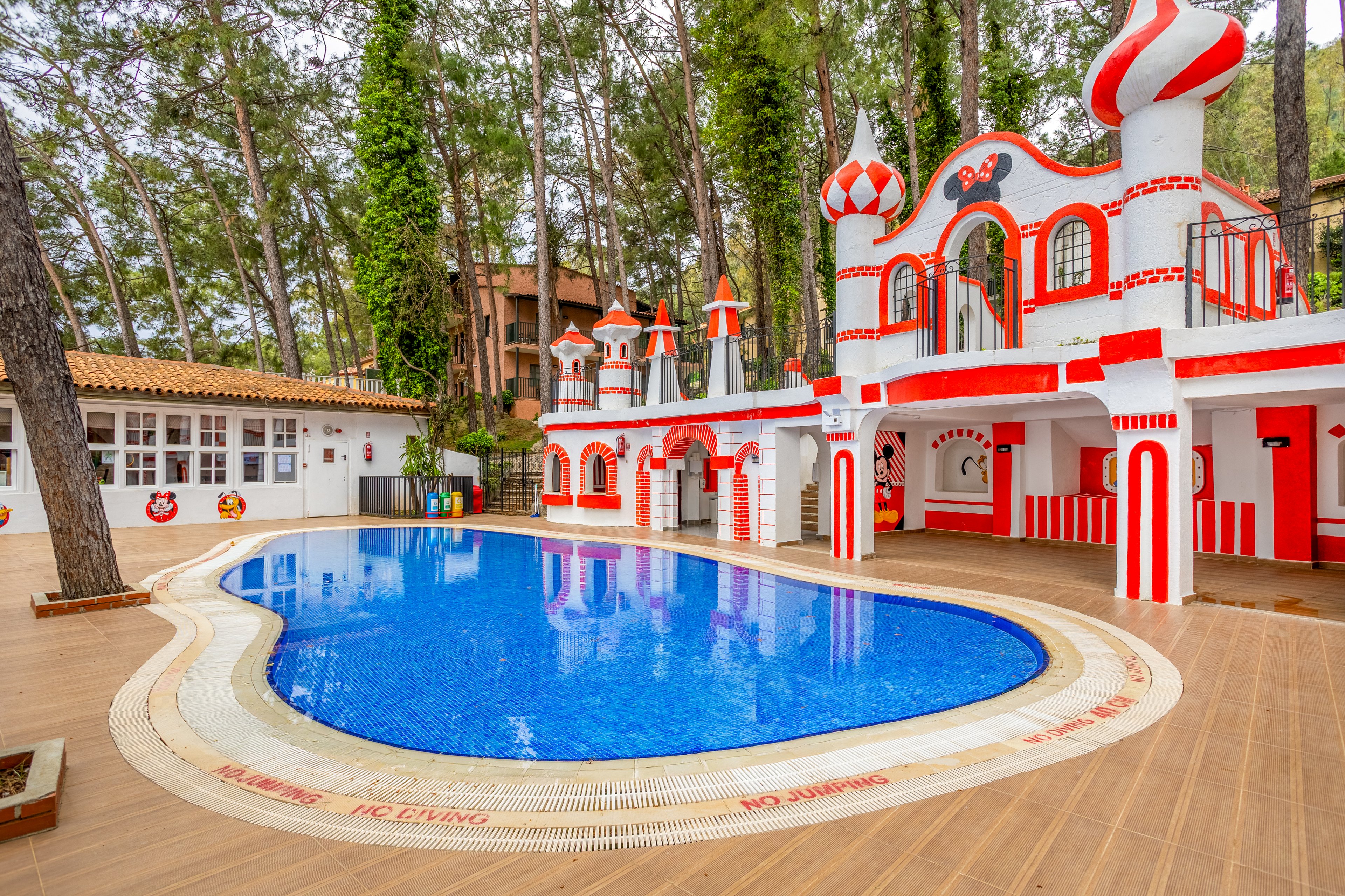 An outdoor swimming pool curves in front of a whimsical red and white castle-like building. The bright blue water contrasts with the warm wooden deck and surrounding pine trees. Playful turrets, arches, and rounded shapes give the structure a fairytale, amusement-park feel. The area appears quiet and empty, suggesting a peaceful resort or themed recreation space in a forest setting.