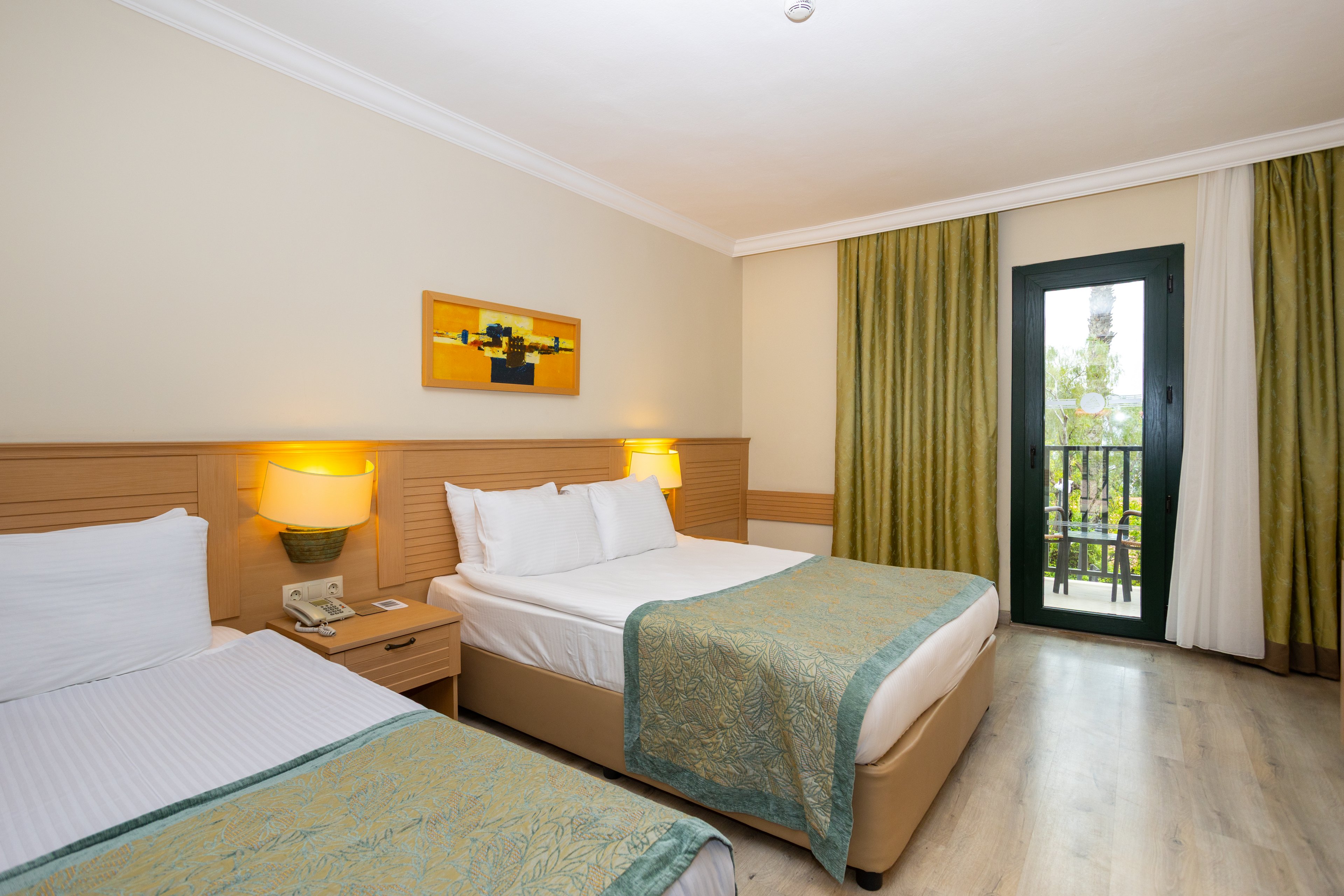 Well-lit hotel room featuring a double bed and a single bed with white linens and green patterned throws. The space includes wooden bedside tables with lamps and a telephone. A large window with green curtains opens to a balcony overlooking greenery. Light-colored walls and flooring create a clean, modern, and inviting atmosphere.