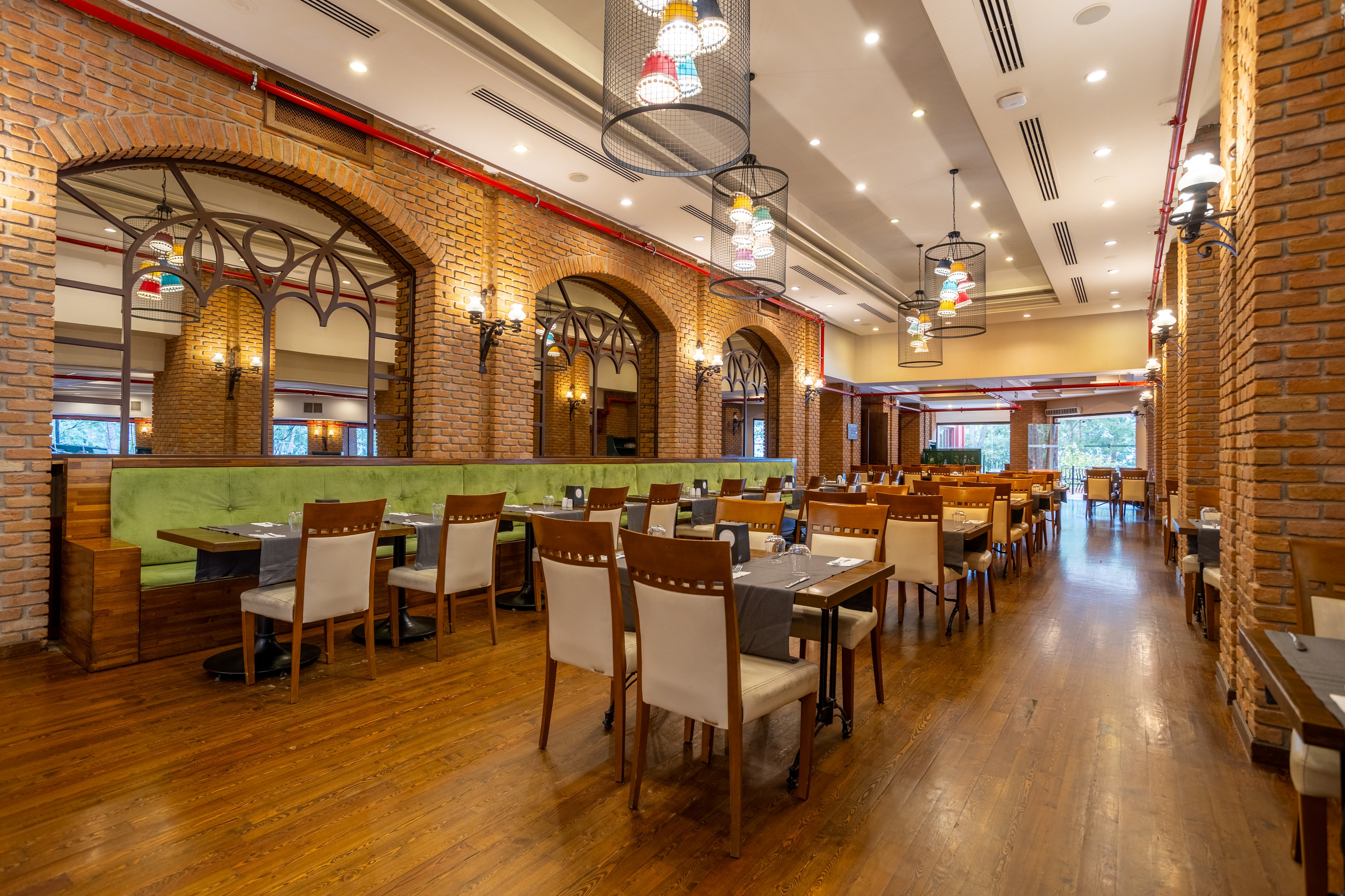 Wide view of a spacious restaurant interior featuring rows of wooden tables and chairs. The setting includes exposed brick walls, arched window frames, and green upholstered booth seating. Warm pendant lights and wall sconces illuminate the room, highlighting the wooden floor and high ceiling. The restaurant appears empty and ready for guests, creating a calm and inviting dining atmosphere.