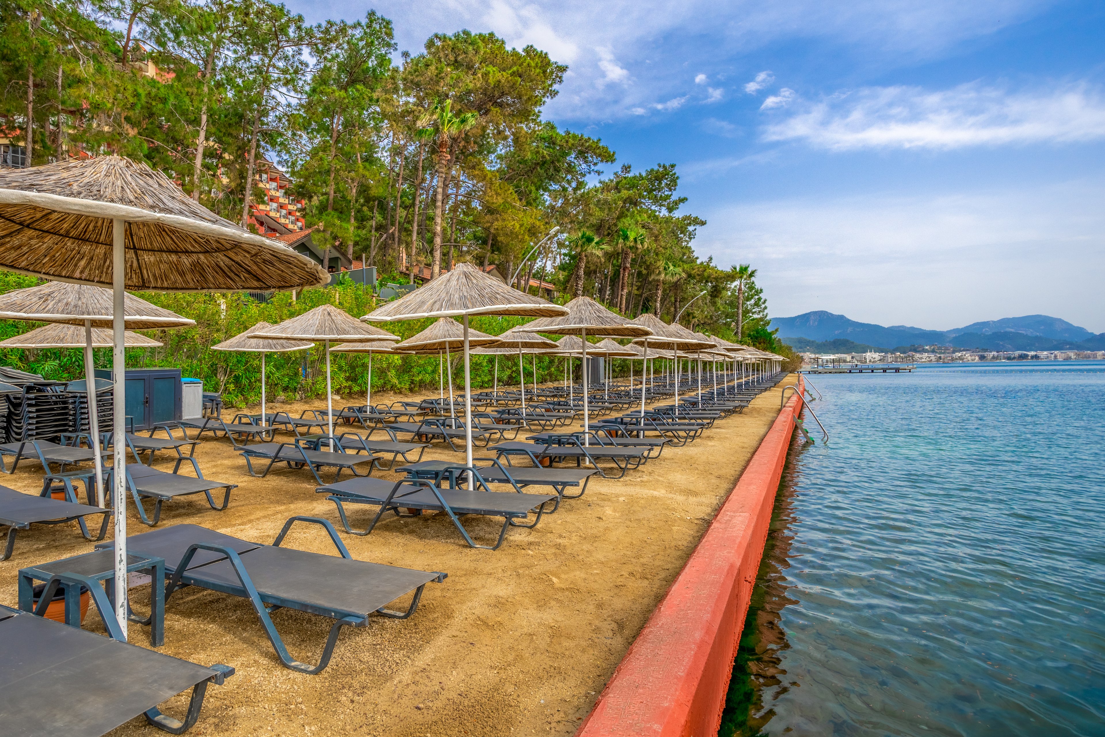 A row of empty sun loungers with straw umbrellas lines a sandy waterfront promenade. The beach area is bordered by a low red wall next to calm, clear blue water. Green trees and vegetation rise behind the loungers, adding a natural backdrop. Distant hills and a partly cloudy sky complete the tranquil coastal scene.