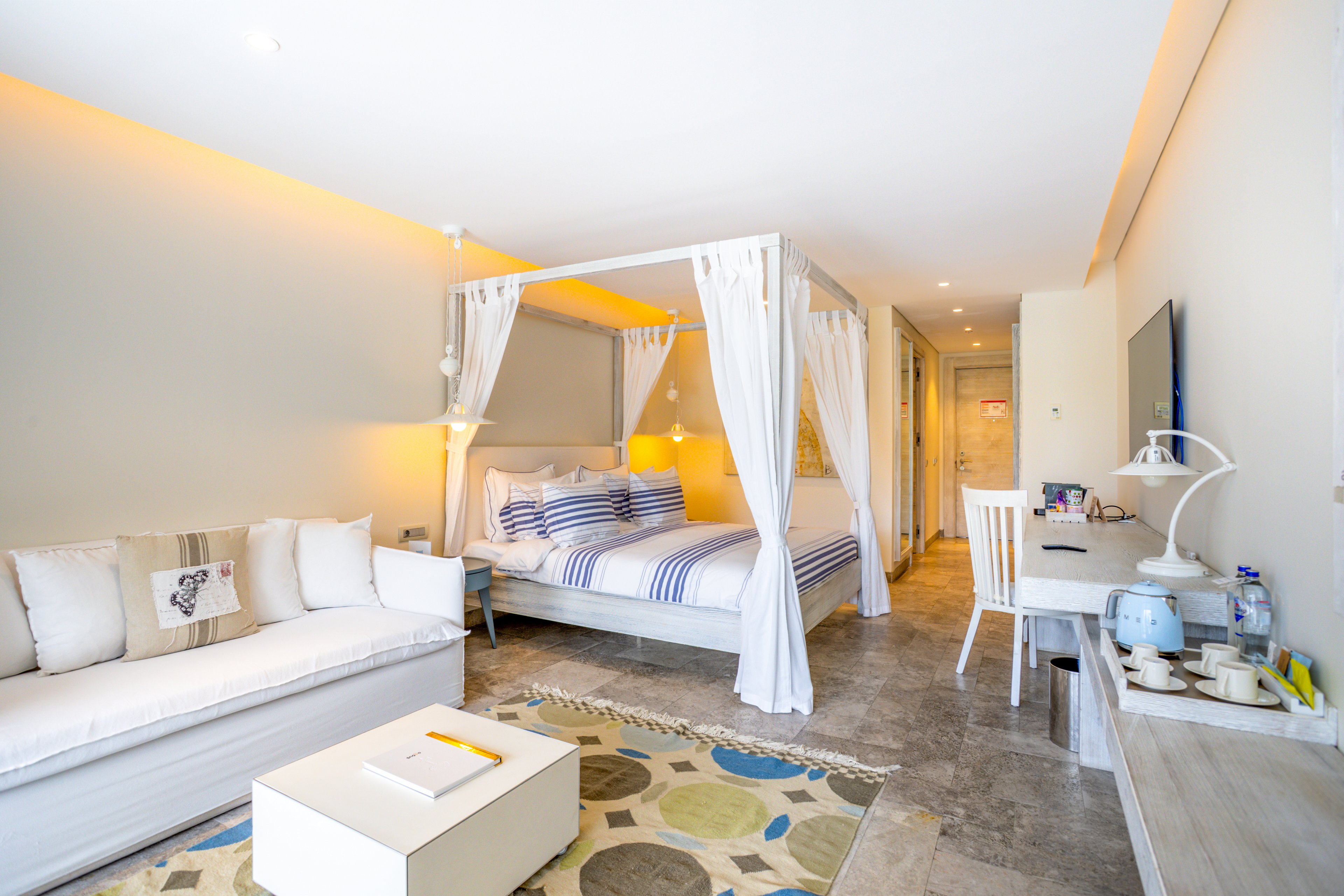 Spacious hotel room featuring a white four-poster canopy bed with blue striped bedding. The setting includes a white sofa, a patterned rug, and a long desk with a flat-screen TV and electric kettle. Warm ambient lighting and light-colored walls create an airy, inviting atmosphere. The room appears tidy and modern, ideal for vacation or resort accommodation.