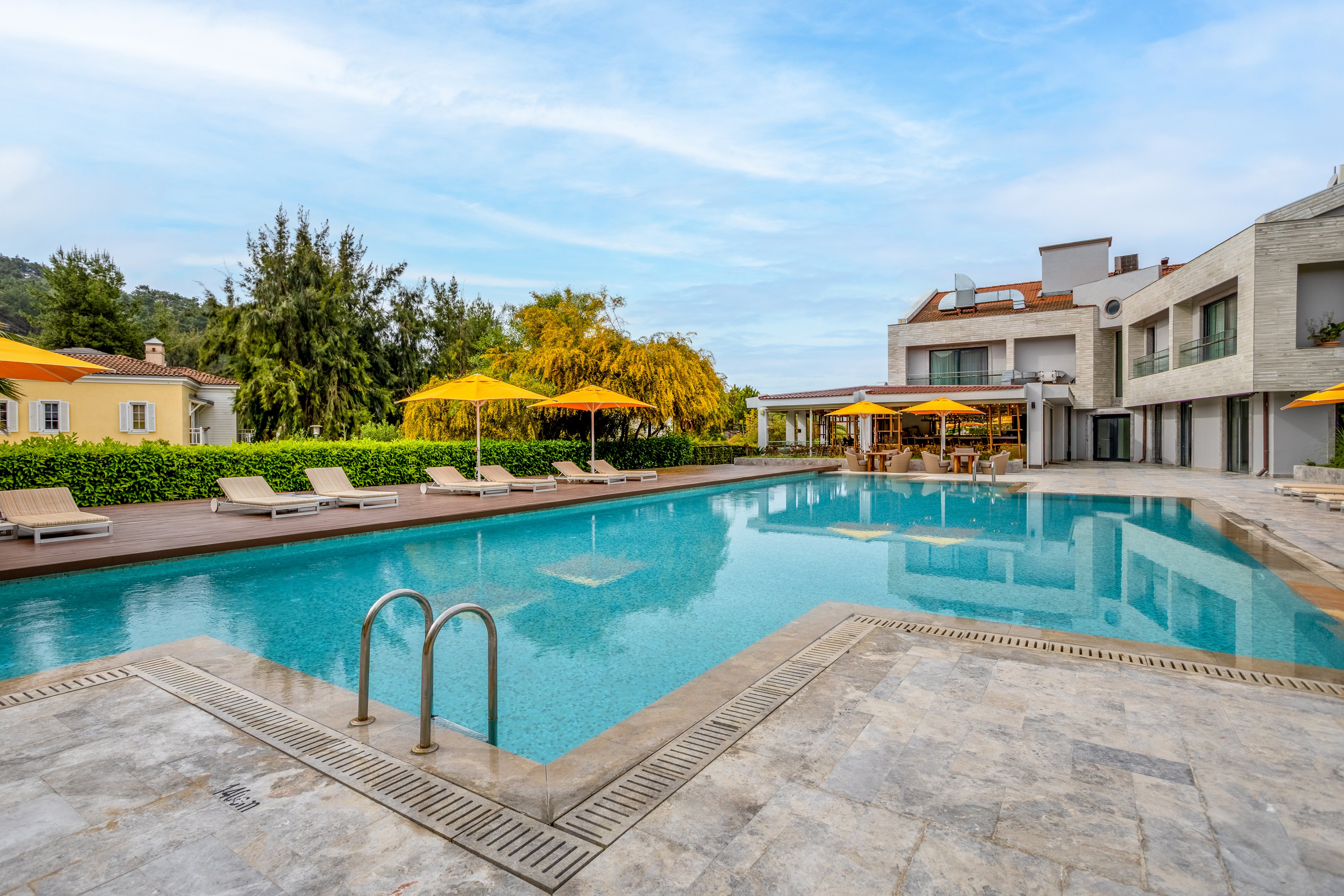 A large outdoor swimming pool sits in front of a modern villa with clean architectural lines. Sun loungers and bright yellow umbrellas line the poolside terrace, creating a relaxing resort atmosphere. The water reflects the surrounding buildings, greenery, and blue sky. Trees and hedges frame the property, suggesting a private and tranquil vacation setting.
