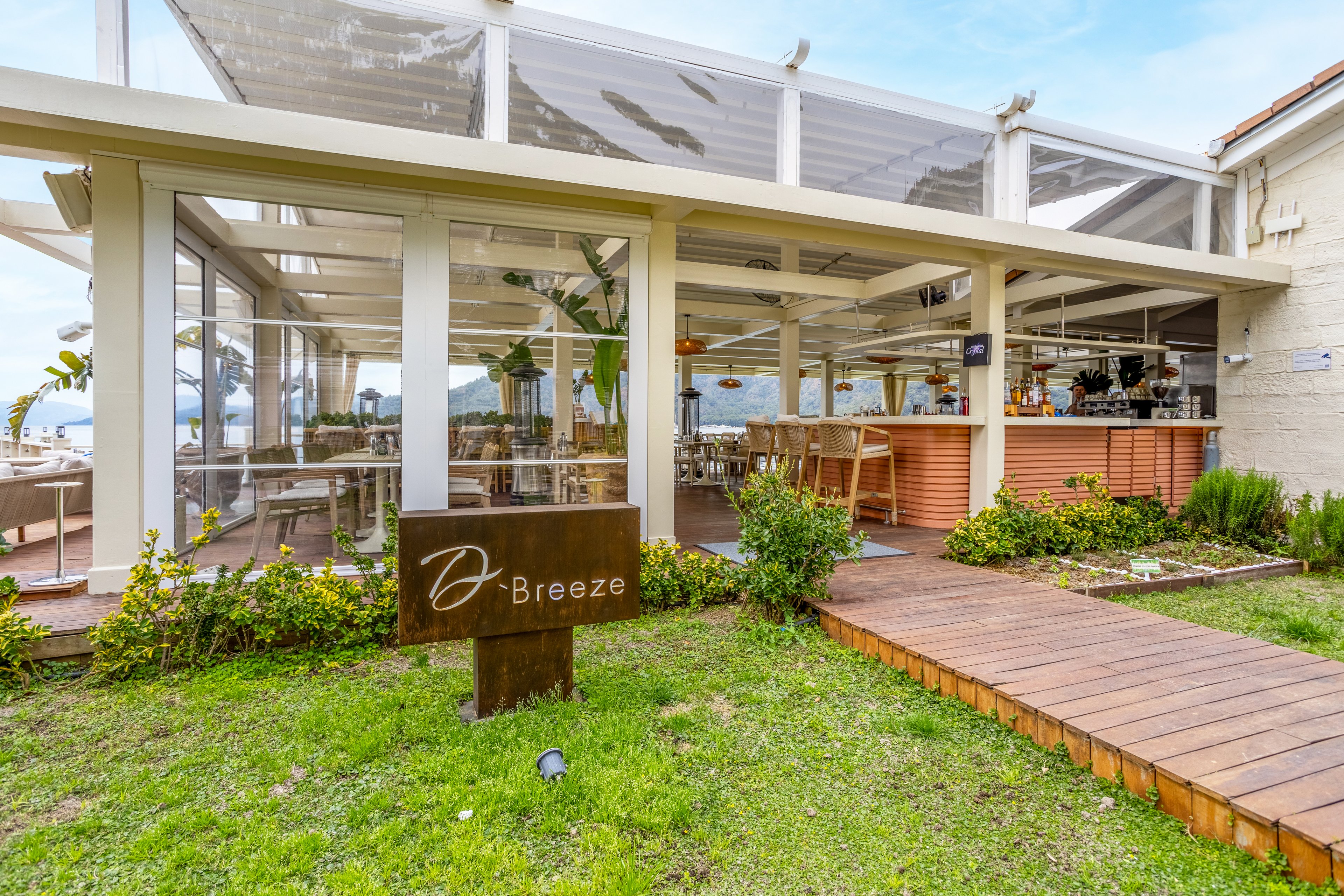 A contemporary seaside restaurant features a glass-enclosed terrace with views of the water. The exterior showcases white structural beams, clear panels, and a wooden walkway leading to the entrance. A metal sign in the foreground reads "Breeze" with a stylized initial above it. Lush green grass and shrubs soften the modern architecture, while the interior bar and seating area appear bright and inviting.