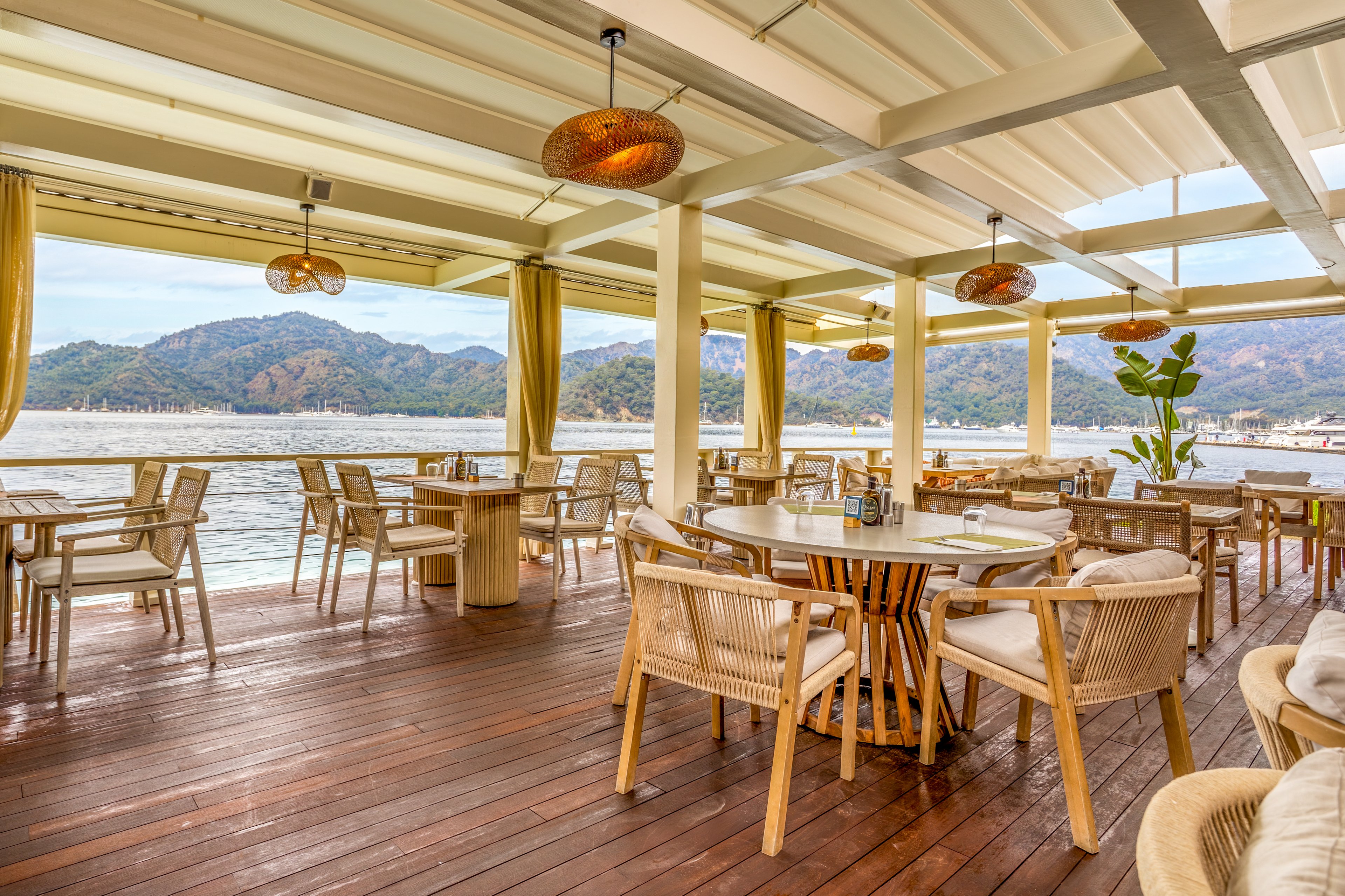 An open-air restaurant terrace overlooks a calm waterfront with distant mountains. Wooden tables and woven chairs are arranged neatly on a polished wooden deck under a covered pergola. Warm pendant lights and soft yellow curtains create a relaxed, coastal ambiance. The scene is bright and inviting, suggesting a tranquil dining experience by the water.