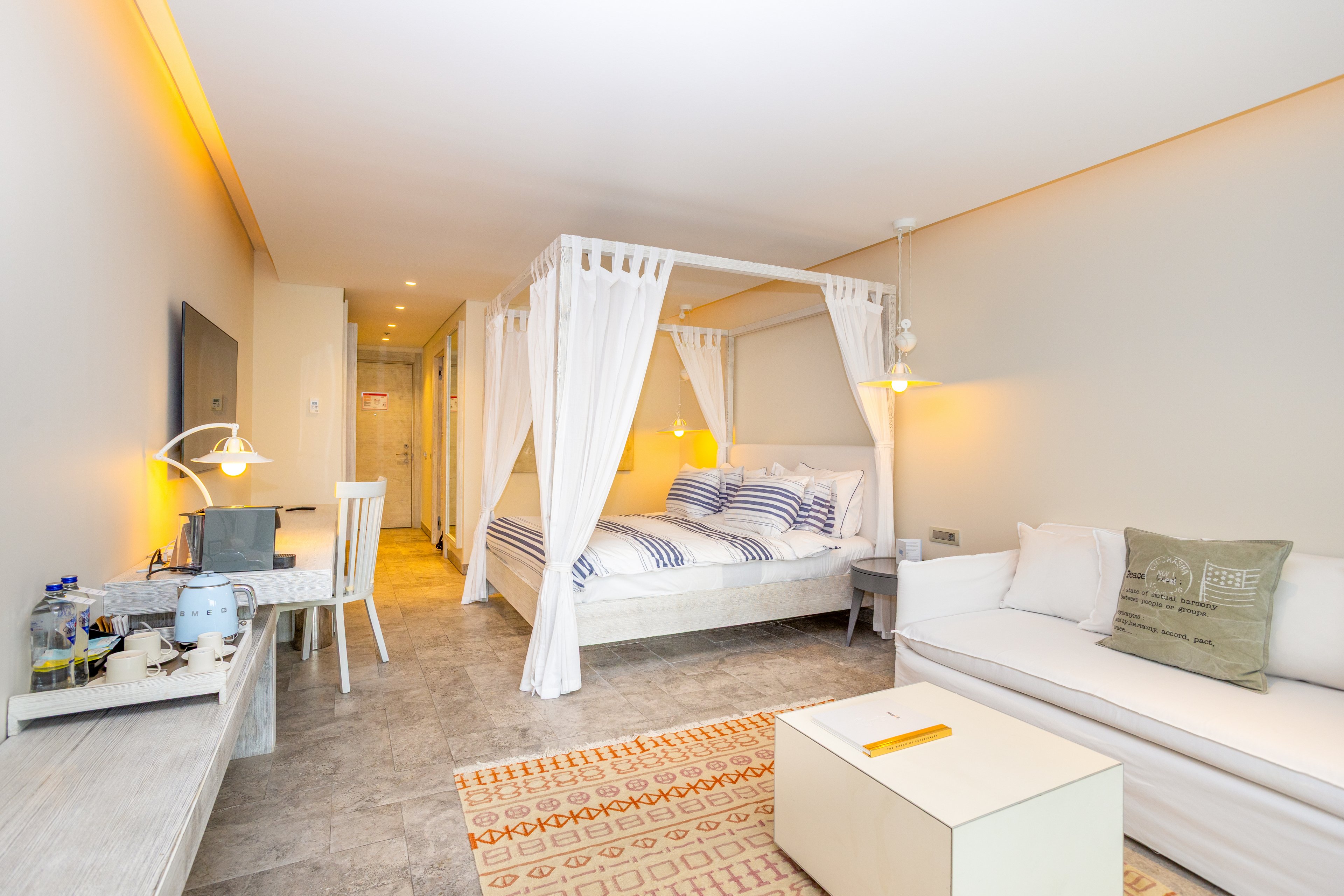 A spacious, modern hotel room features a white four-poster canopy bed with striped bedding as the focal point. The room includes a white sofa, a work desk with a lamp, and a flat-screen TV mounted on the wall. Warm recessed lighting and neutral walls create a cozy, inviting atmosphere. A patterned rug and light-colored flooring add texture and contrast to the clean, minimalist decor.