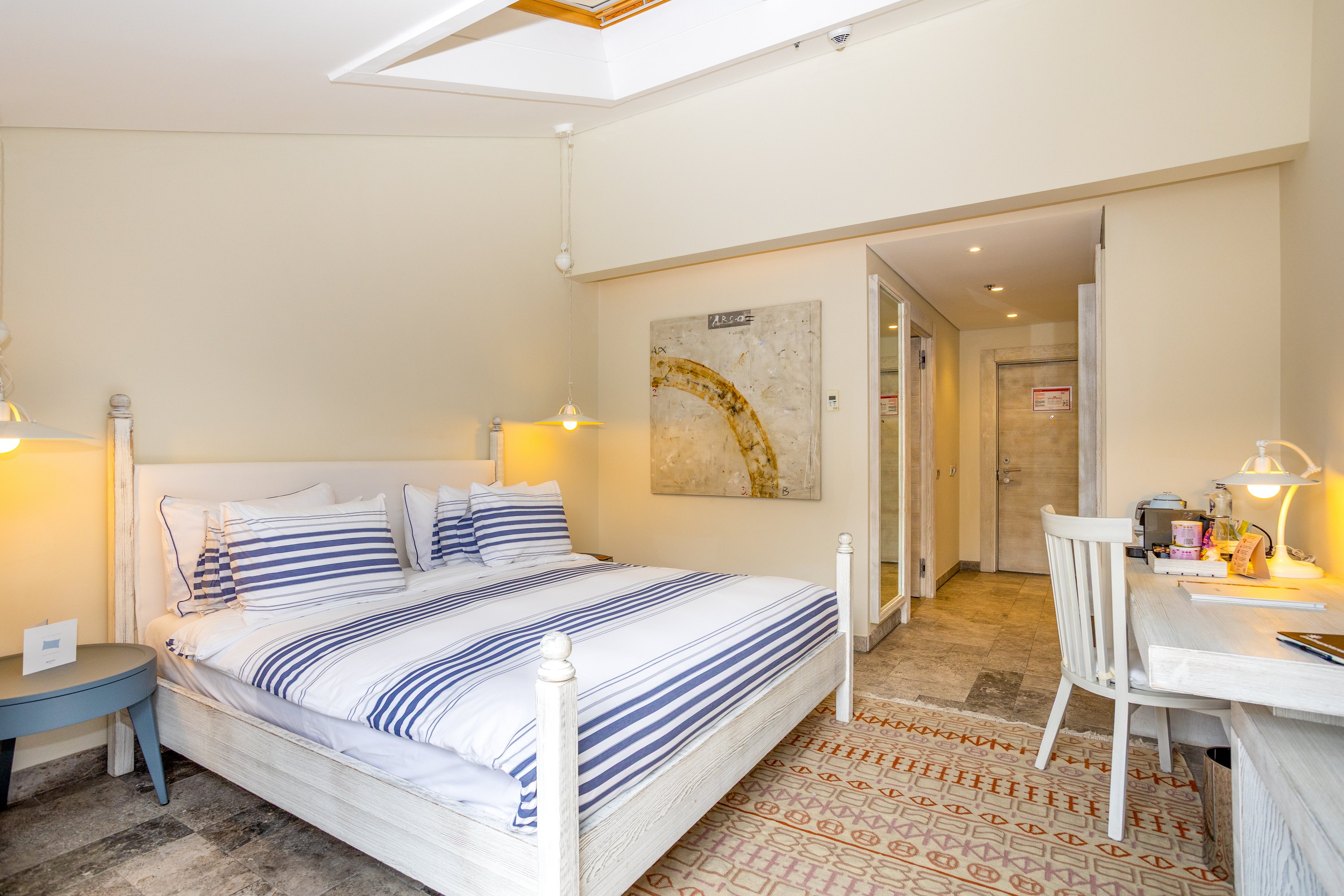 A neatly made double bed with blue and white striped bedding is the focal point of this bright hotel room. The space features light-colored walls, a patterned rug, and a small round side table next to the bed. A desk with a white chair, lamp, and various items provides a functional workspace along one wall. Recessed lighting and a skylight-style ceiling detail add to the airy, contemporary feel of the room.