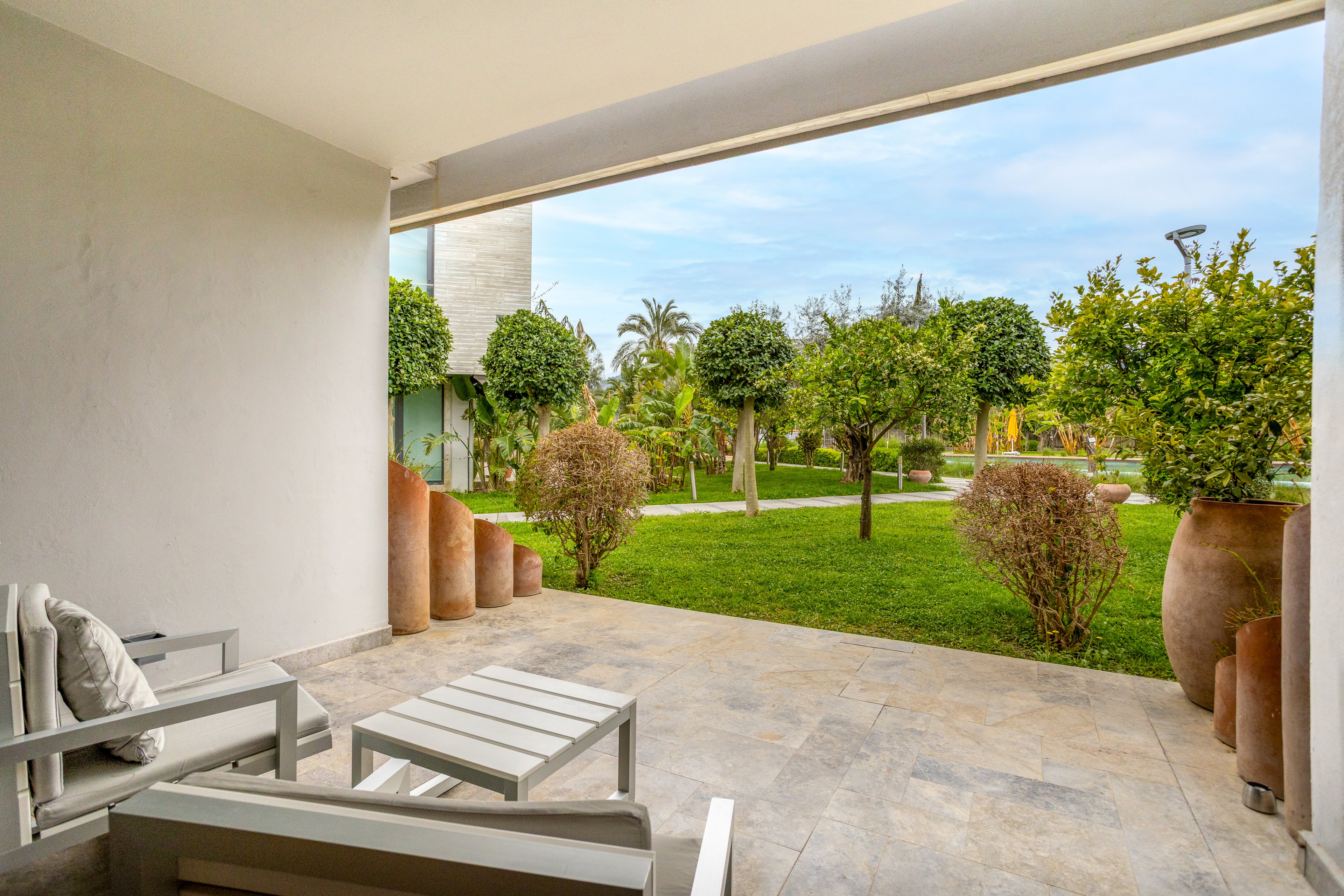 A modern covered patio features minimalist white lounge chairs and a small table on a stone floor. The terrace opens onto a well-maintained green lawn with neatly trimmed trees and shrubs. Large decorative clay pots line the edge of the patio, adding a natural design element. In the background, contemporary buildings and palm trees suggest a resort or residential complex under a bright, clear sky.