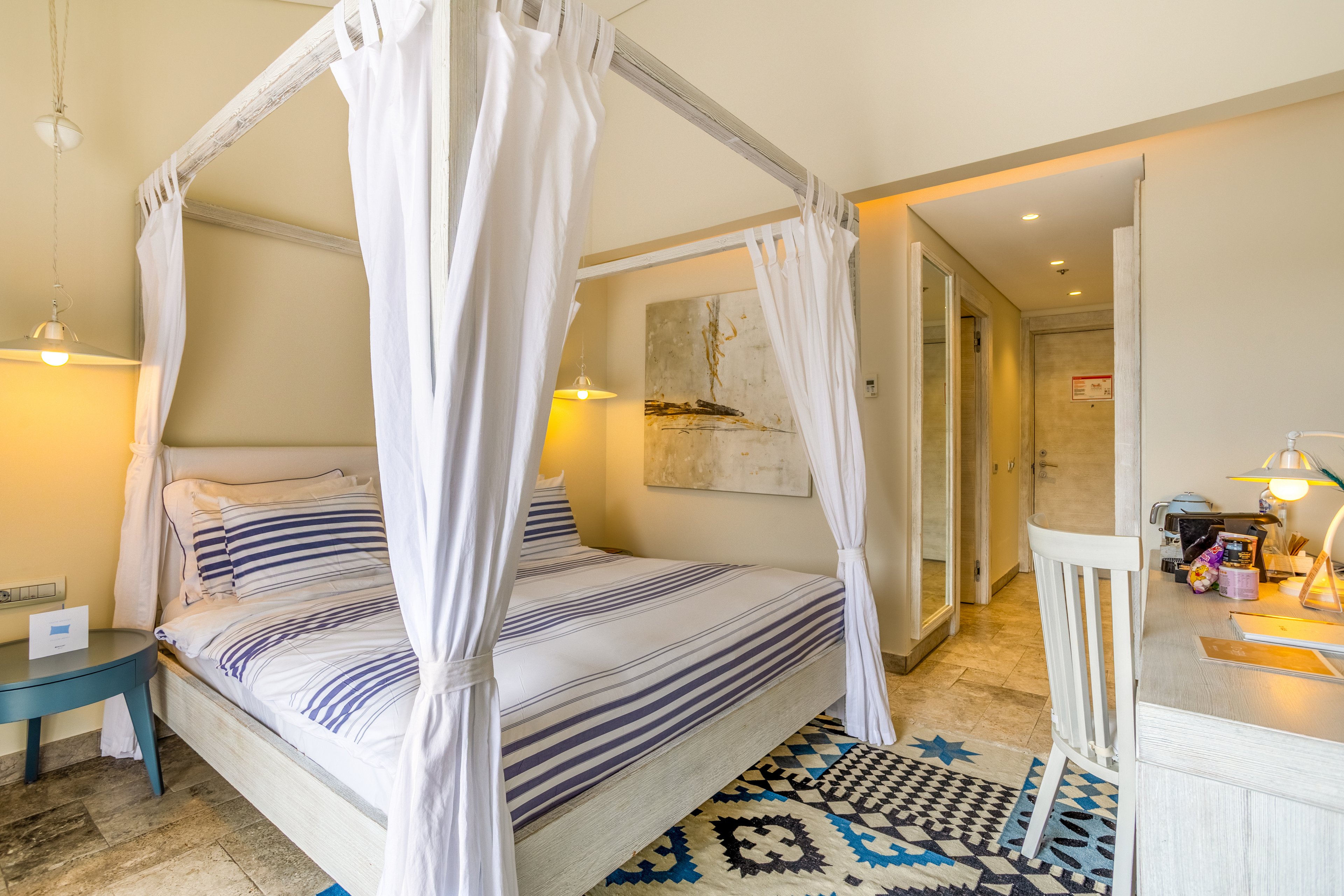 A cozy bedroom features a white four-poster canopy bed dressed in blue and white striped bedding. The room is decorated in a light coastal style with warm beige walls and a patterned rug in blue, black, and white tones. A small bedside table with a lamp and a desk with a chair and workspace accessories sit along the walls. Soft lighting and natural tones create an inviting, relaxing atmosphere in this modern interior.