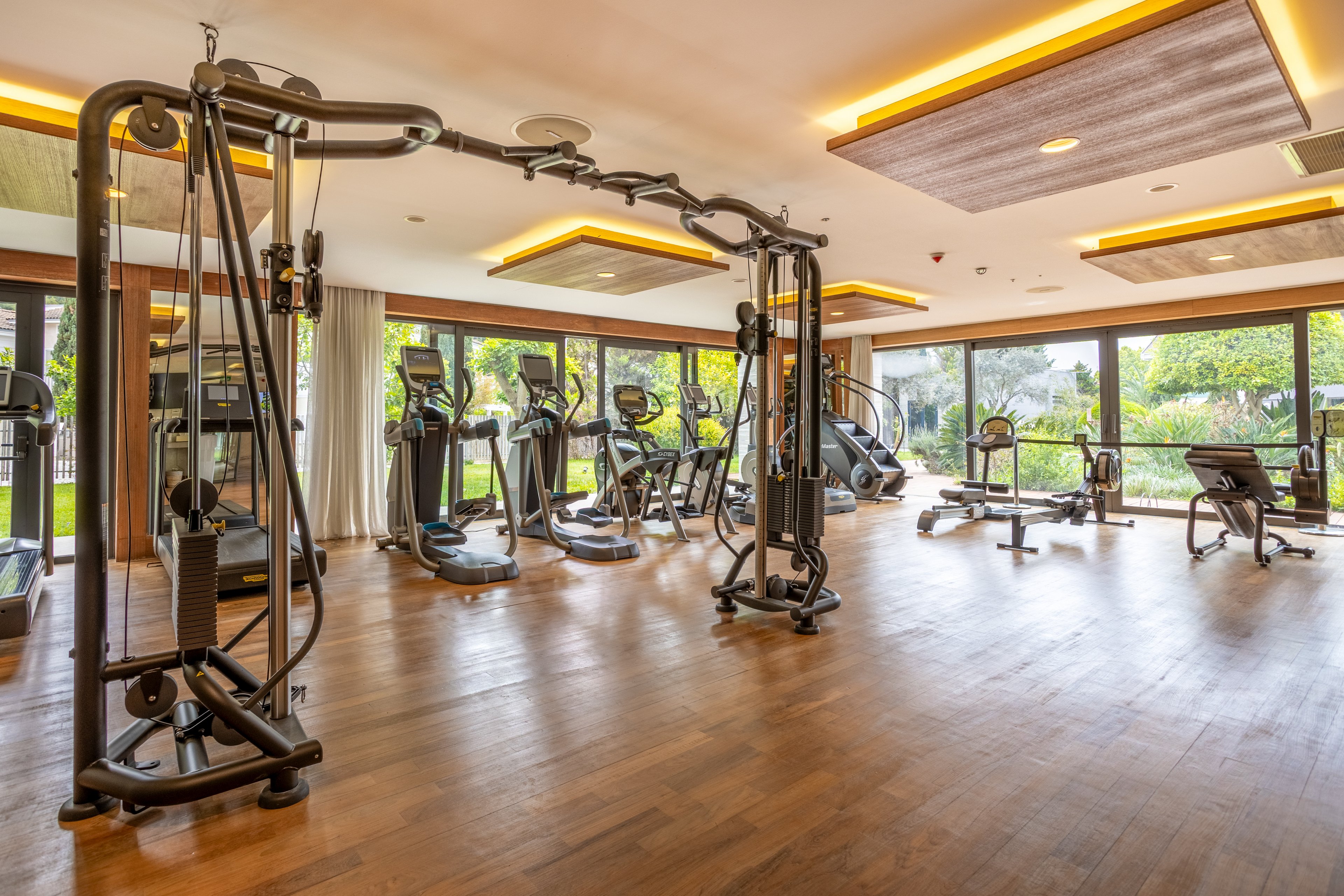 A spacious, modern fitness center features a variety of exercise machines, including a large cable station, treadmills, and stationary bikes. The gym has warm wooden flooring and recessed ceiling lighting that creates an inviting atmosphere. Floor-to-ceiling windows line one side of the room, offering views of greenery and natural light. The space appears clean, organized, and ready for guests to work out.