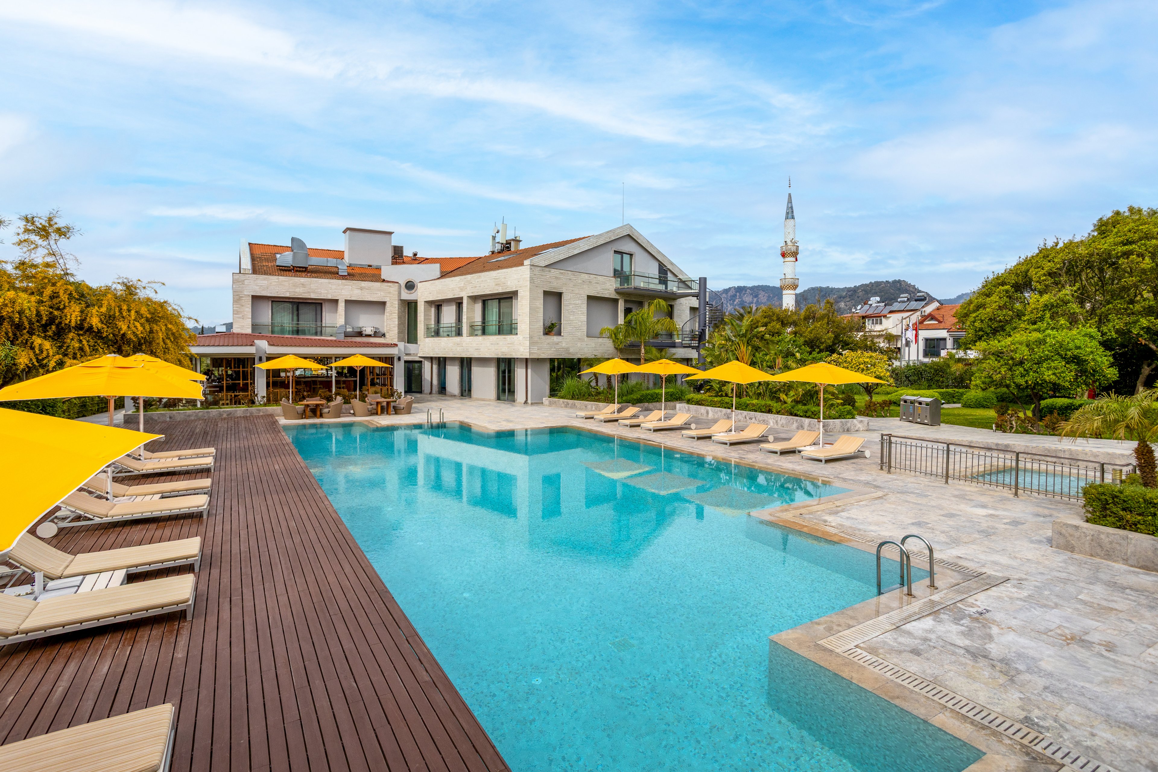 A modern hotel building overlooks a large outdoor swimming pool with clear blue water. Wooden decking and stone tiles surround the pool, lined with beige loungers and bright yellow umbrellas. The scene is set in a landscaped garden with green trees and shrubs, under a blue sky with light clouds. In the background, a tall minaret and nearby houses indicate a town or residential area beyond the hotel grounds.