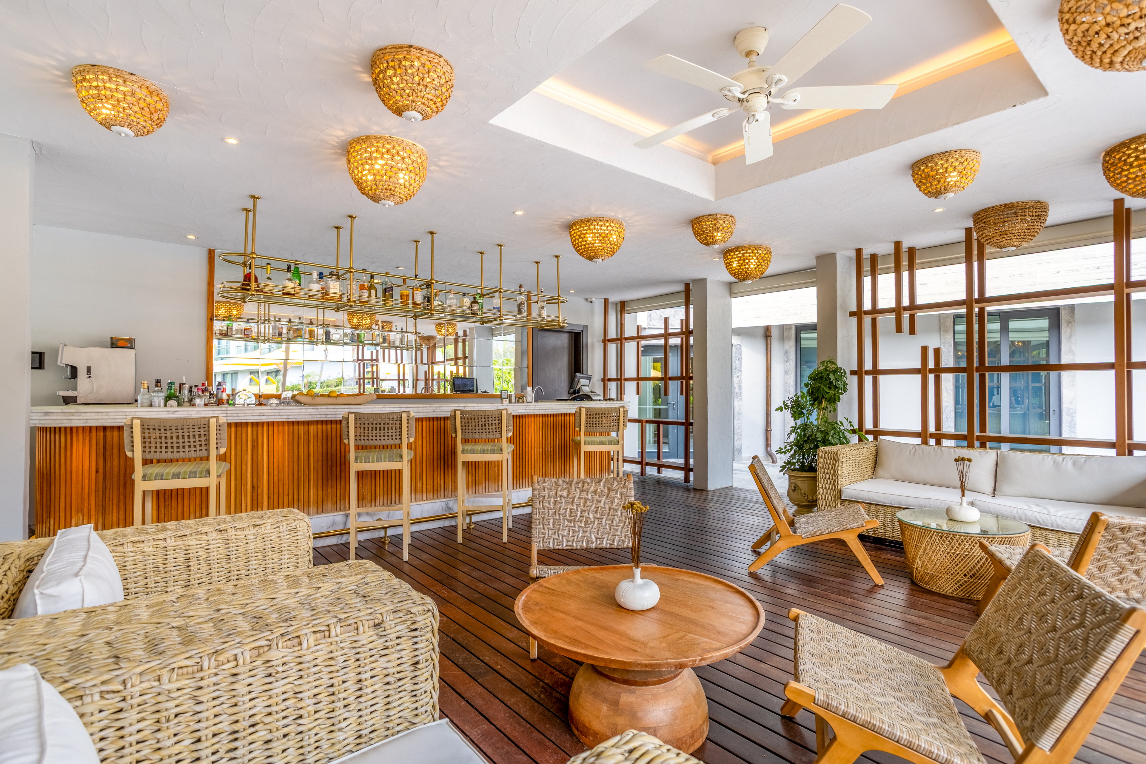 Contemporary lounge bar interior with wicker seating and wooden accents. The space features a long bar counter with high stools and a well-stocked back bar. Large windows and glass doors let in natural light, highlighting the warm wood flooring and white ceiling with woven pendant lights. Neutral-toned sofas and chairs create a relaxed, tropical atmosphere suitable for socializing or casual dining.
